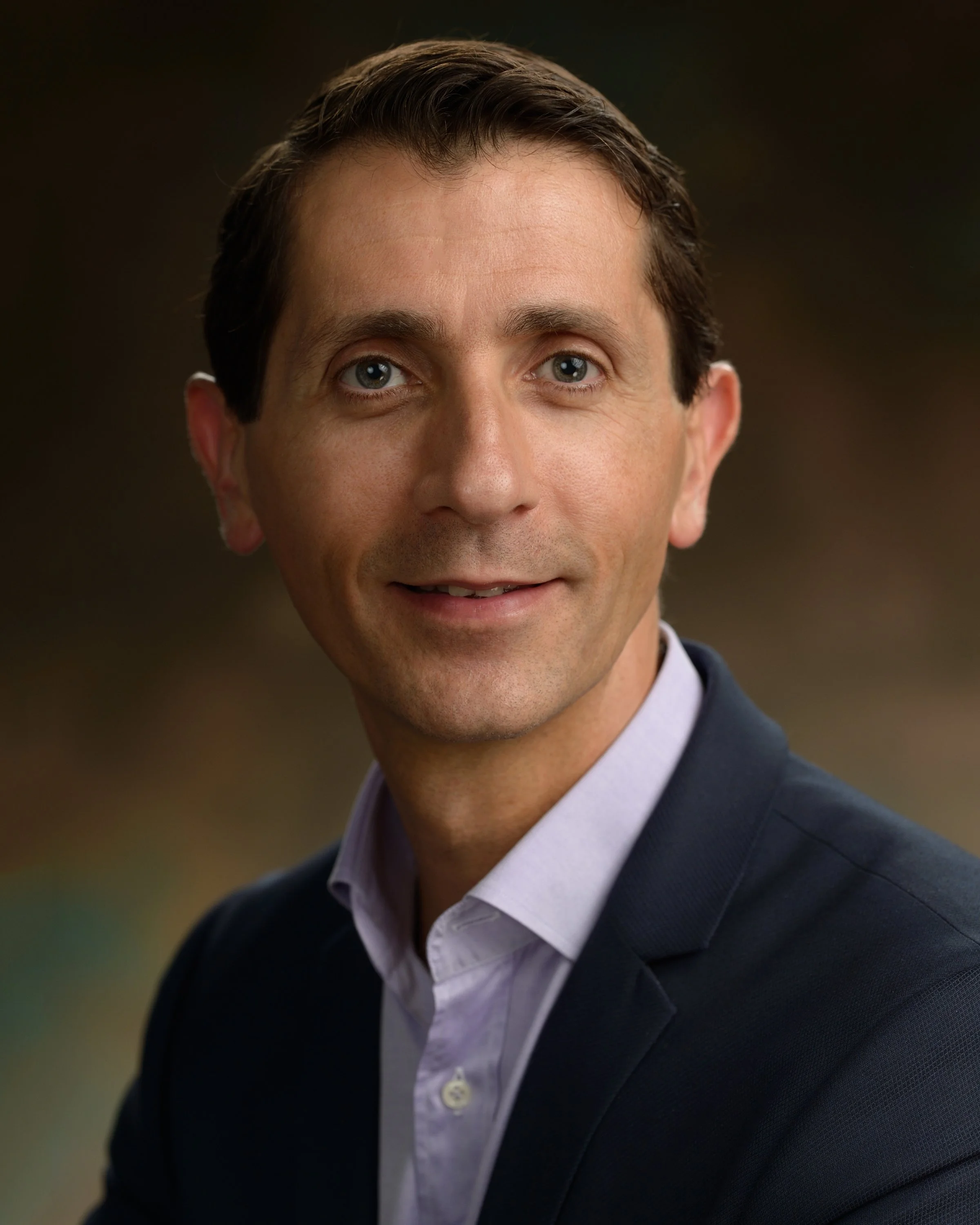 A professional headshot of a man in a dark blazer and light-colored shirt, smiling slightly, with brown hair and blue eyes, set against a blurred background.