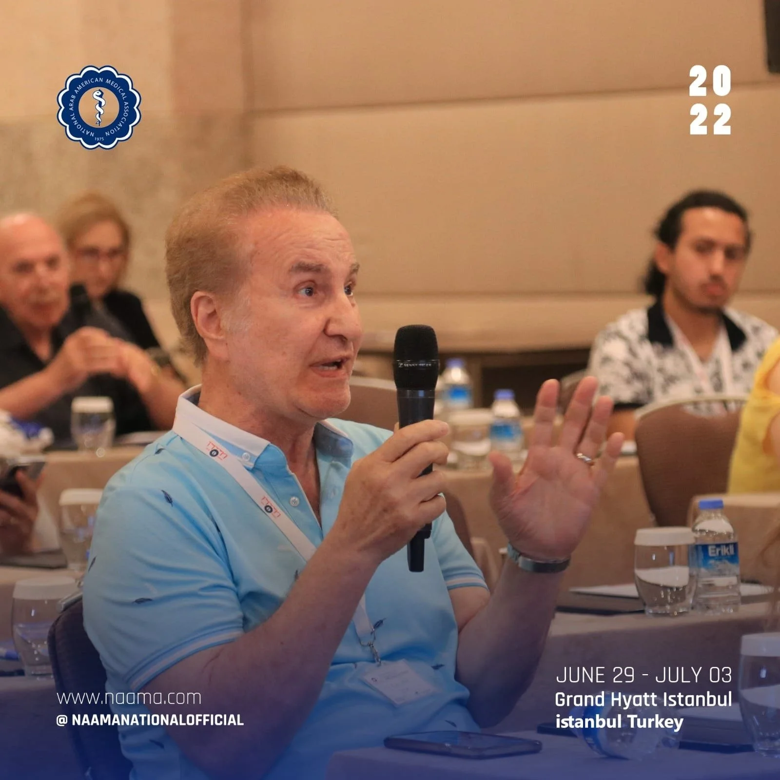 Man with short, light-colored hair and a light blue polo shirt speaking into a microphone at a conference, with other attendees sitting at tables behind him, water bottles on the tables, and a Marriott badge around his neck. The event is in Istanbul,