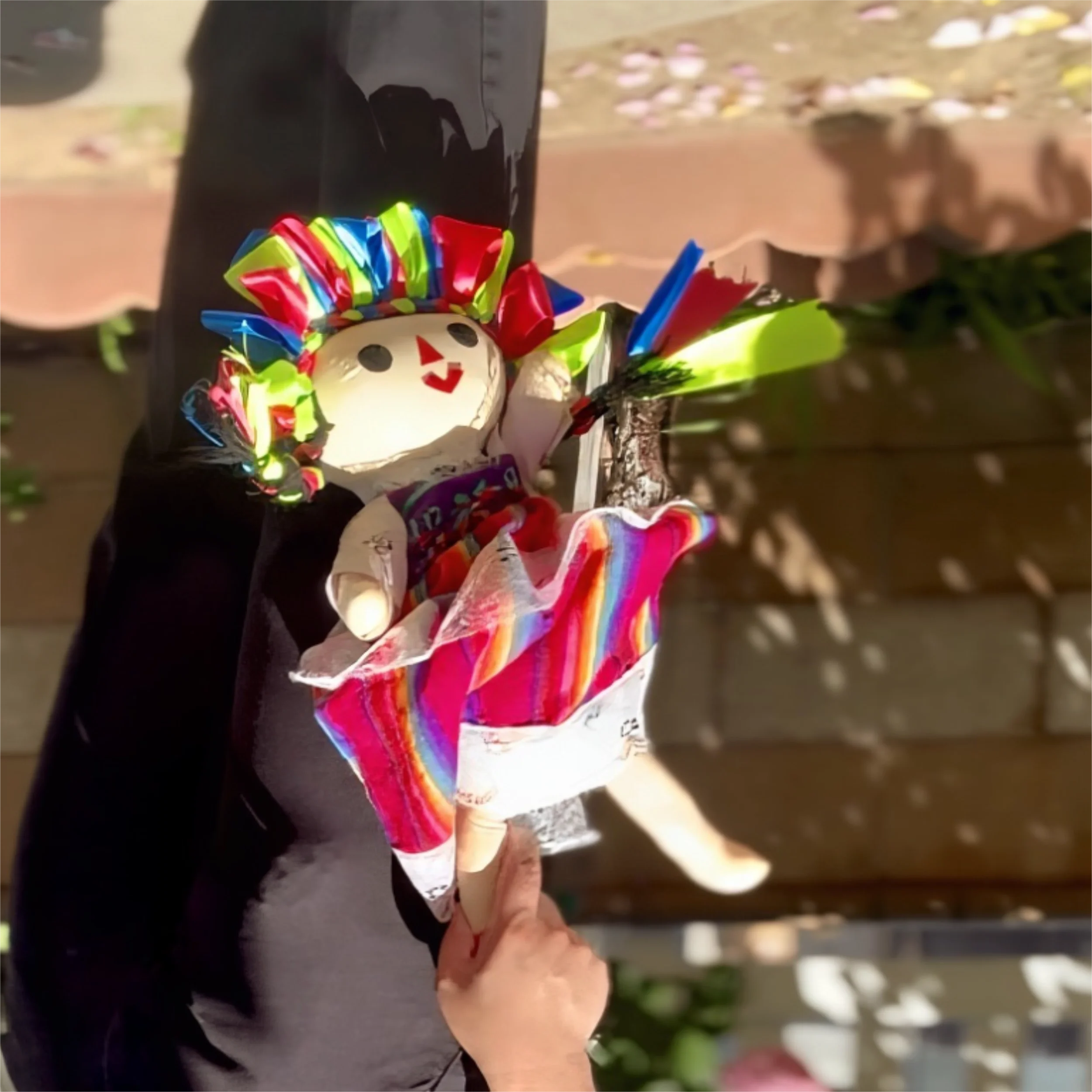 A person holding a small snowman figurine decorated with colorful ribbon hair, a red nose, and a face made of black dots, with a background of falling flower petals.