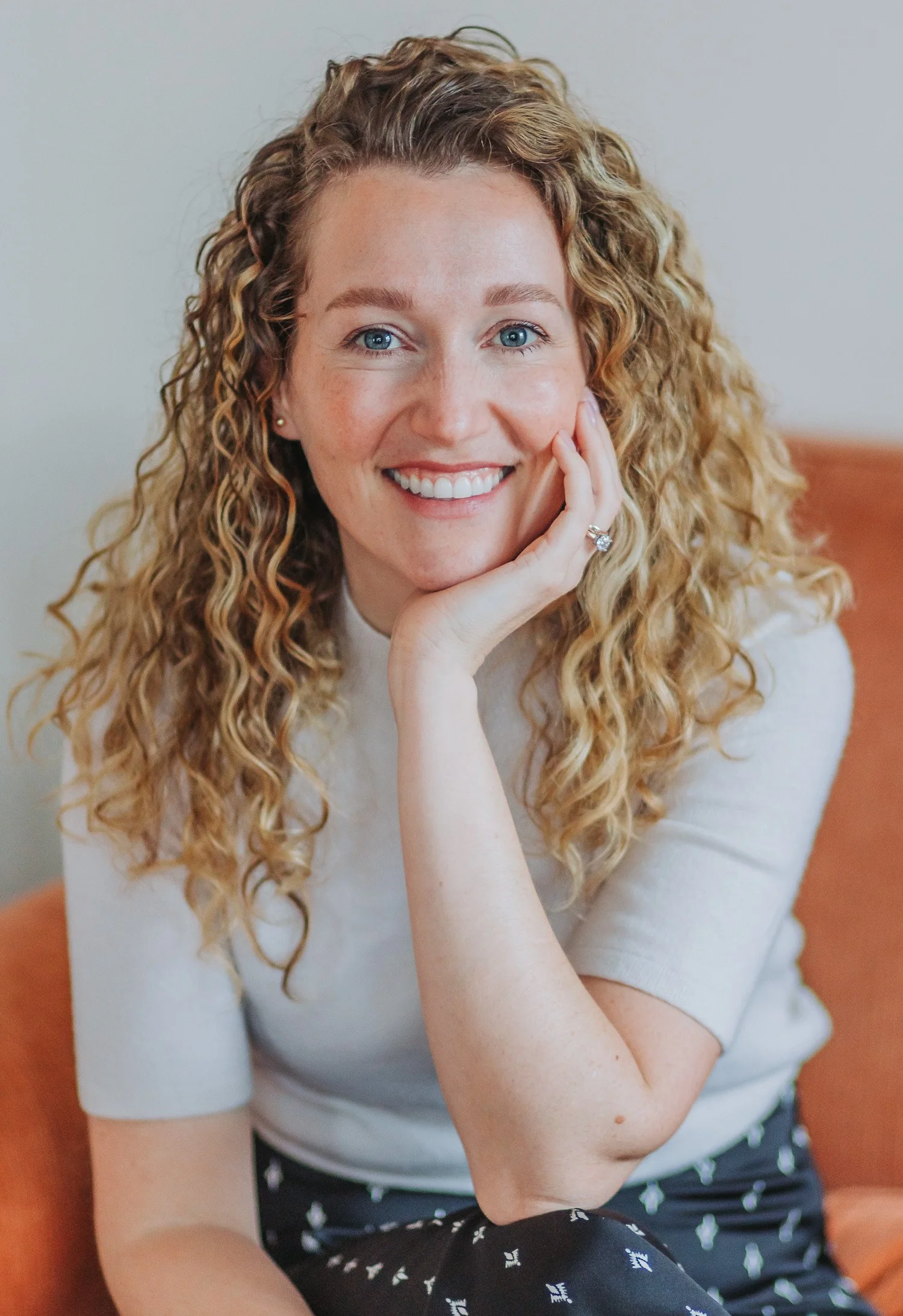 A woman with curly blonde hair, blue eyes, and a bright smile sitting on an orange chair, resting her chin on her hand, wearing a white top and patterned black pants.