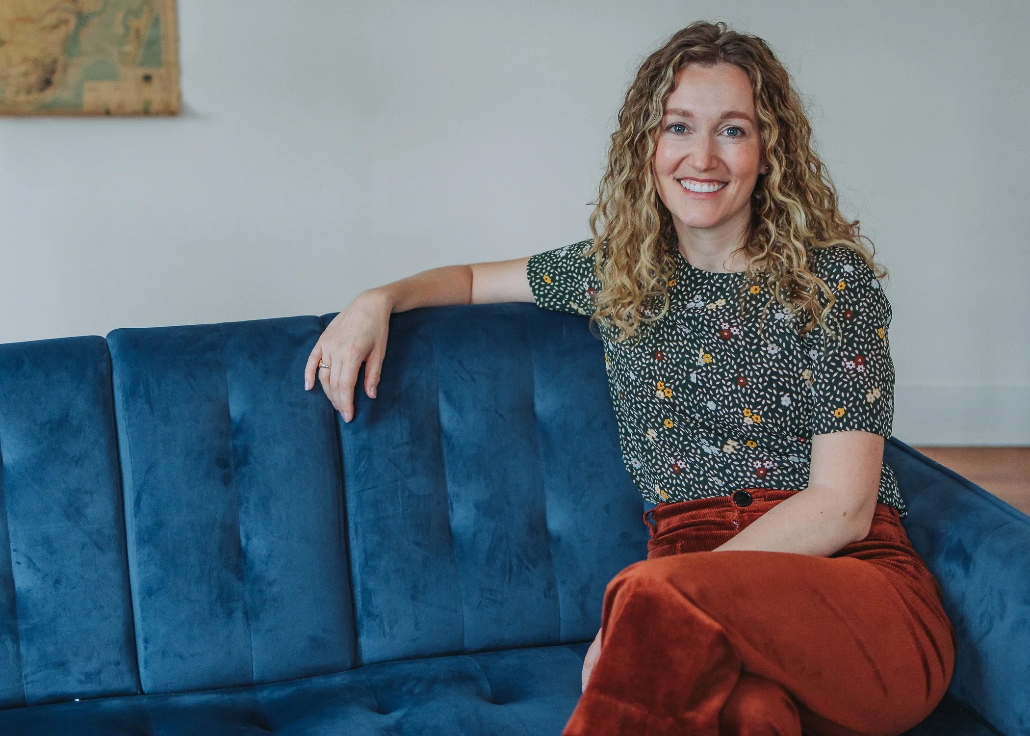 A smiling woman with curly blonde hair sitting on a blue sofa in a room with light-colored walls.
