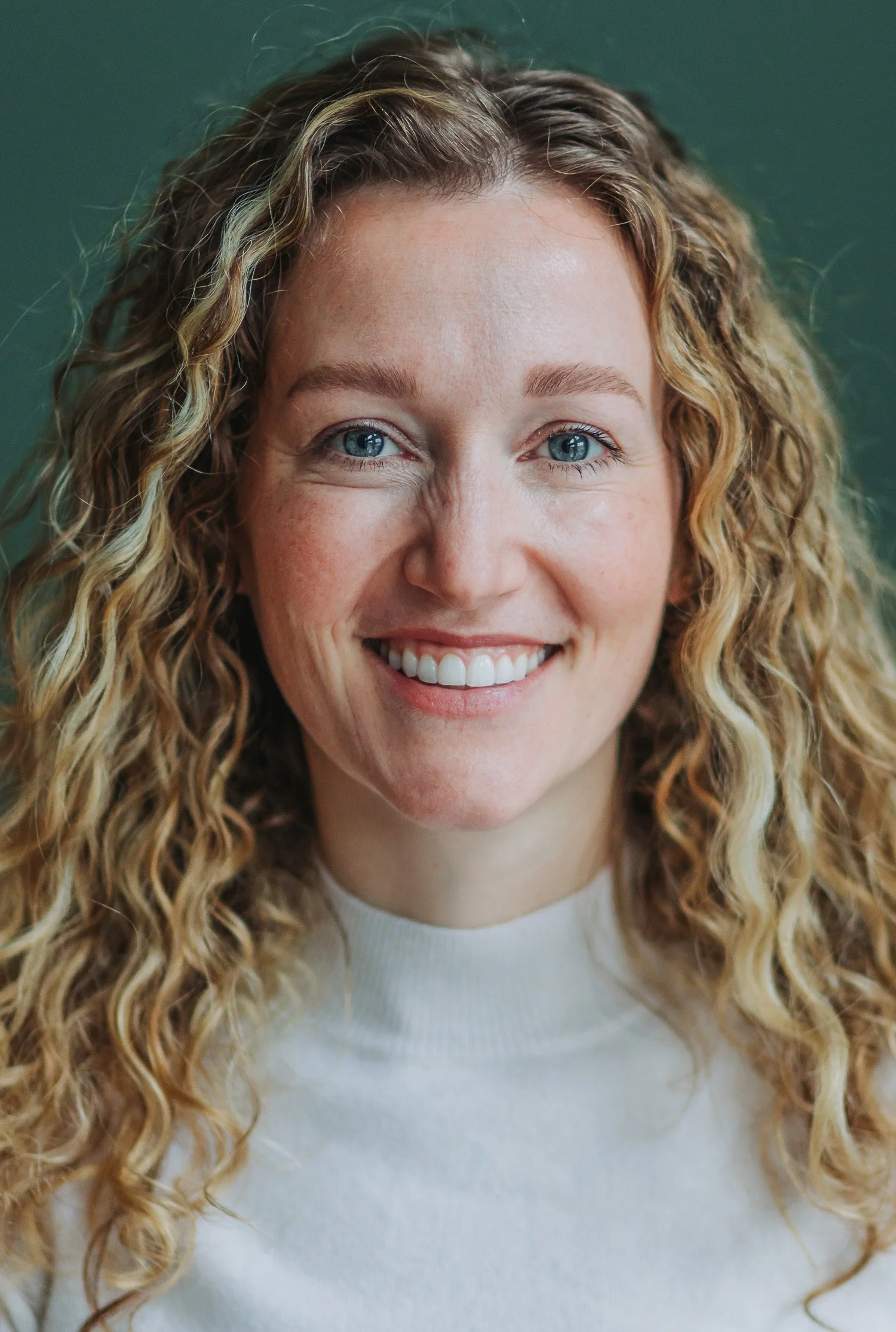 Close-up of a smiling woman with curly blonde hair, blue eyes, and fair skin wearing a white shirt.