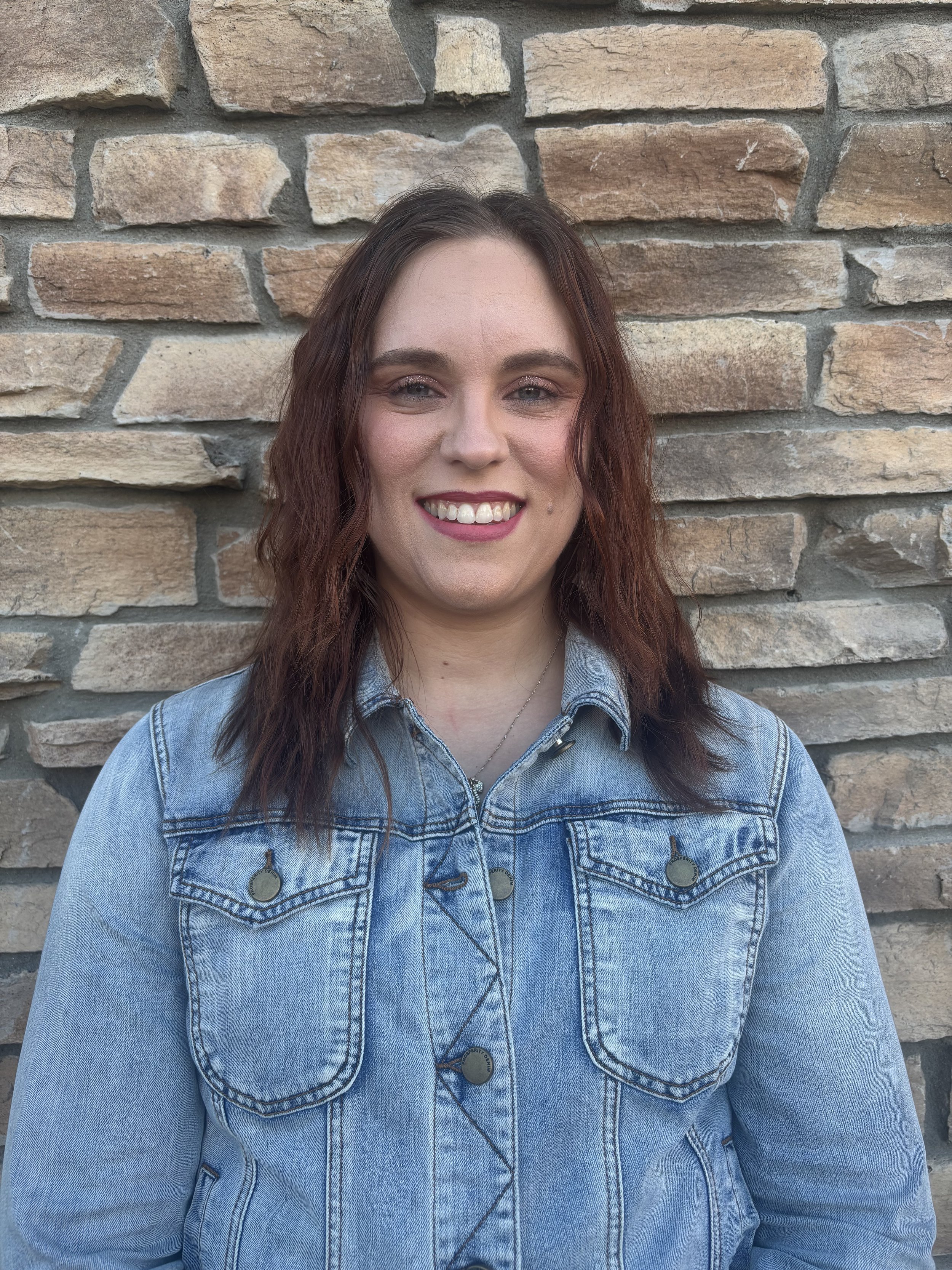 A woman with wavy, reddish-brown hair smiling and standing in front of a brick wall. She is wearing a light blue denim jacket with metal buttons.