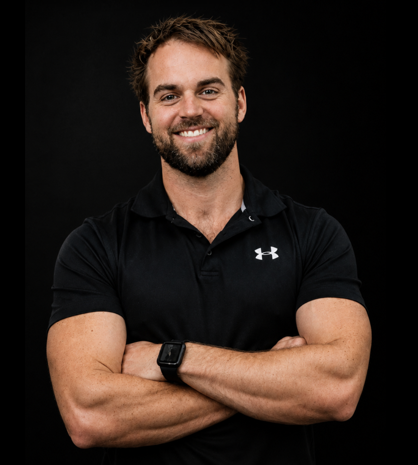 A smiling man with a beard, wearing a black Under Armour polo shirt and a smartwatch, standing with arms crossed against a black background.