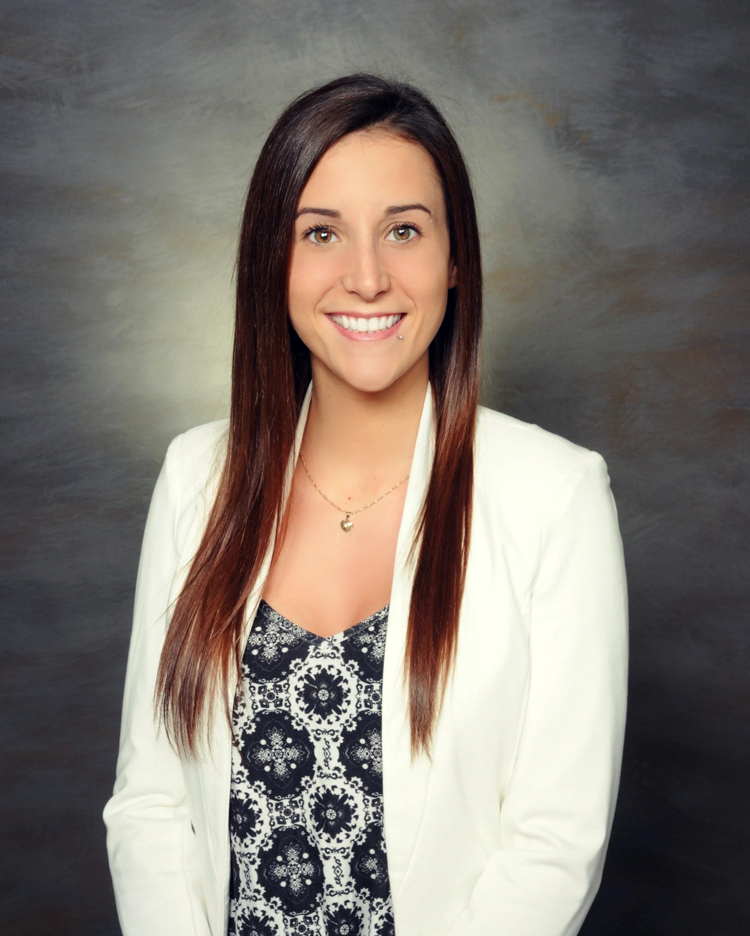 Portrait of a smiling woman with long brown hair, wearing a white blazer, a black and white patterned top, a heart-shaped necklace, and a lip piercing, against a grayish background.