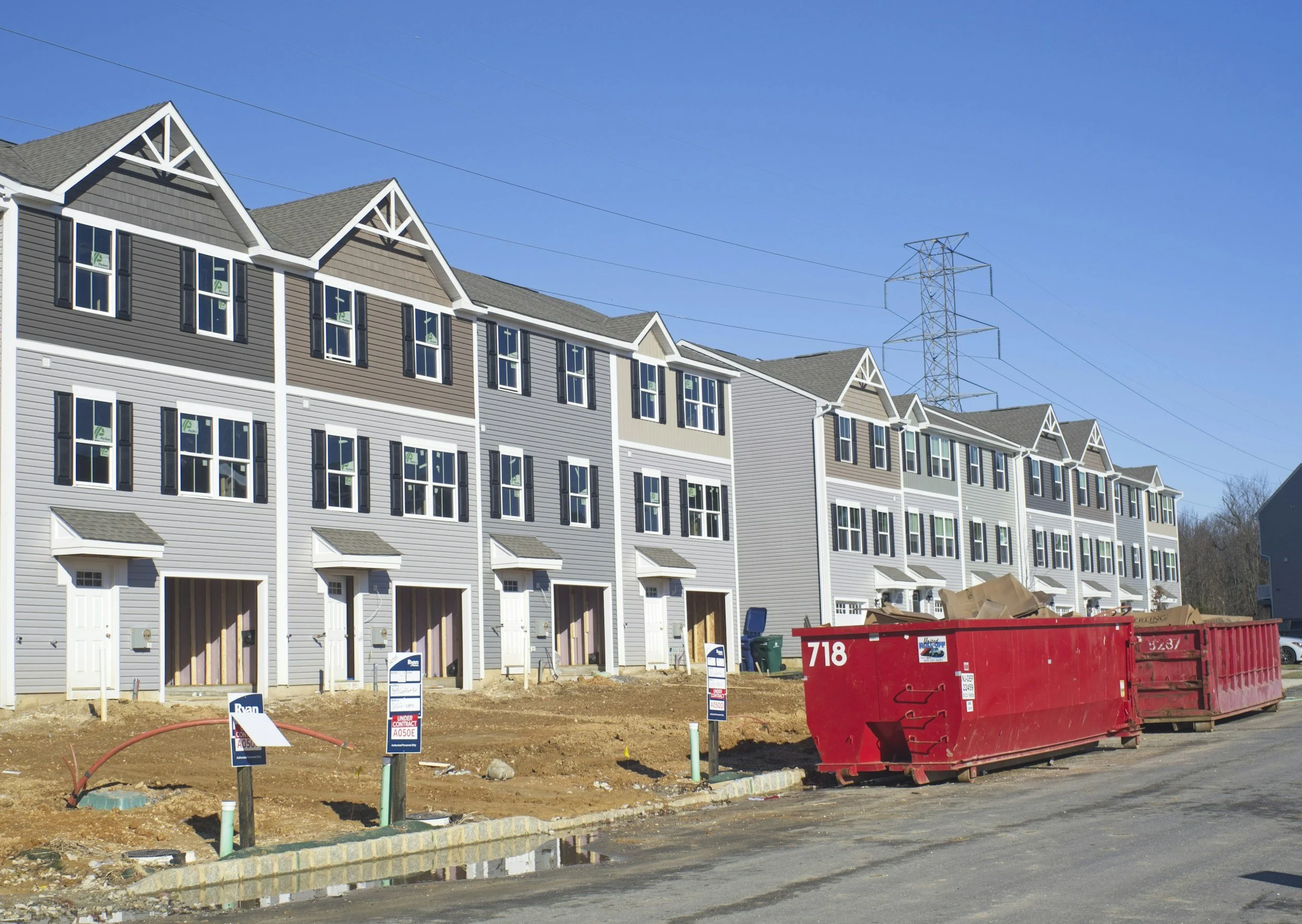Under construction row of modern townhouses with large windows, some with black shutters, and small front porches, against a clear blue sky. Construction signs and a red dumpster are in the foreground.