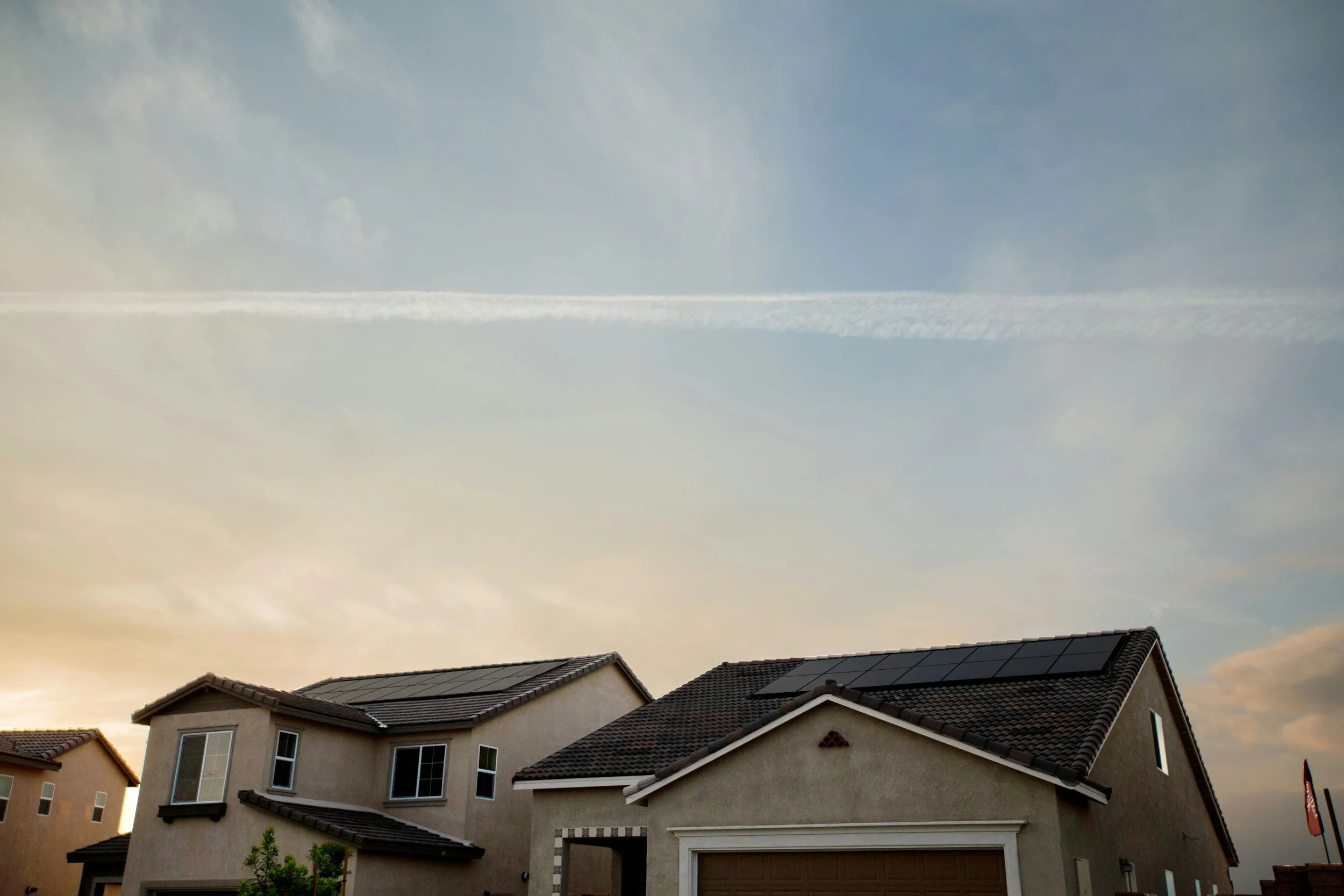 Suburban neighborhood with multiple houses, brown roofs, and a sunset sky with clouds and contrails.