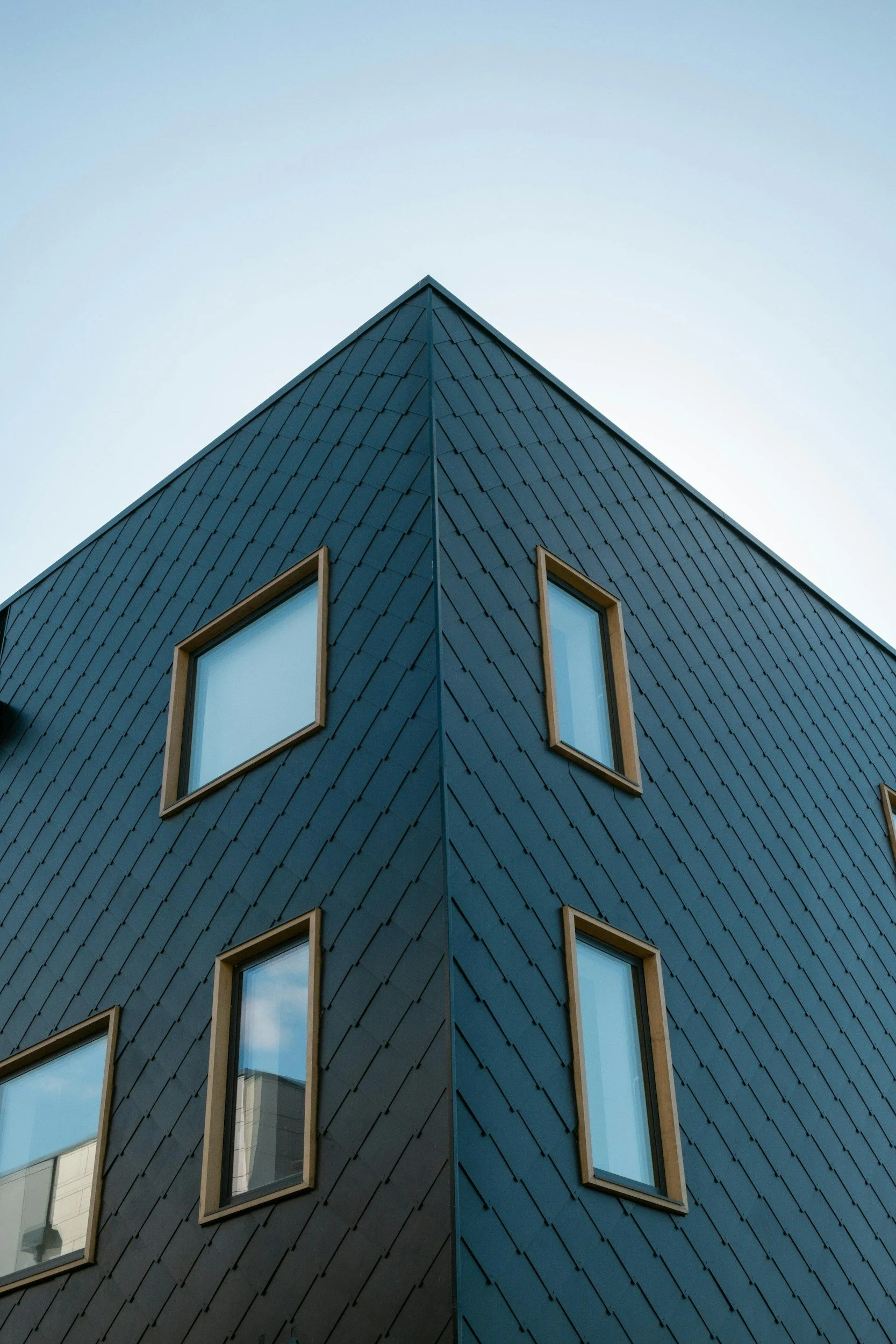 Close-up of a modern building's corner with dark shingle siding, four rectangular windows framed in wood, and a pale sky background.