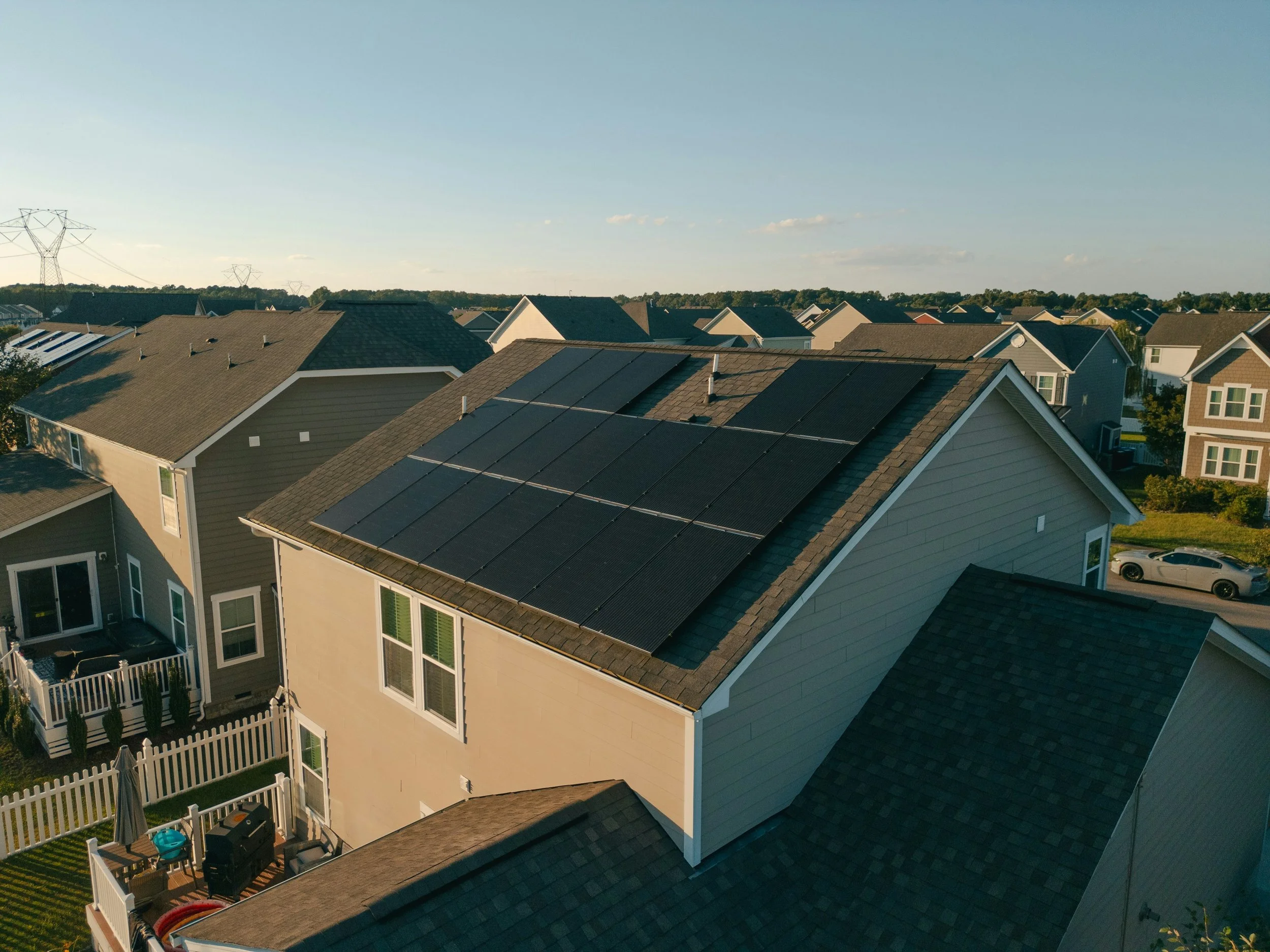 Aerial view of a suburban neighborhood with houses featuring solar panels on their roofs, fences, and a backyard with patio furniture, under a clear blue sky.