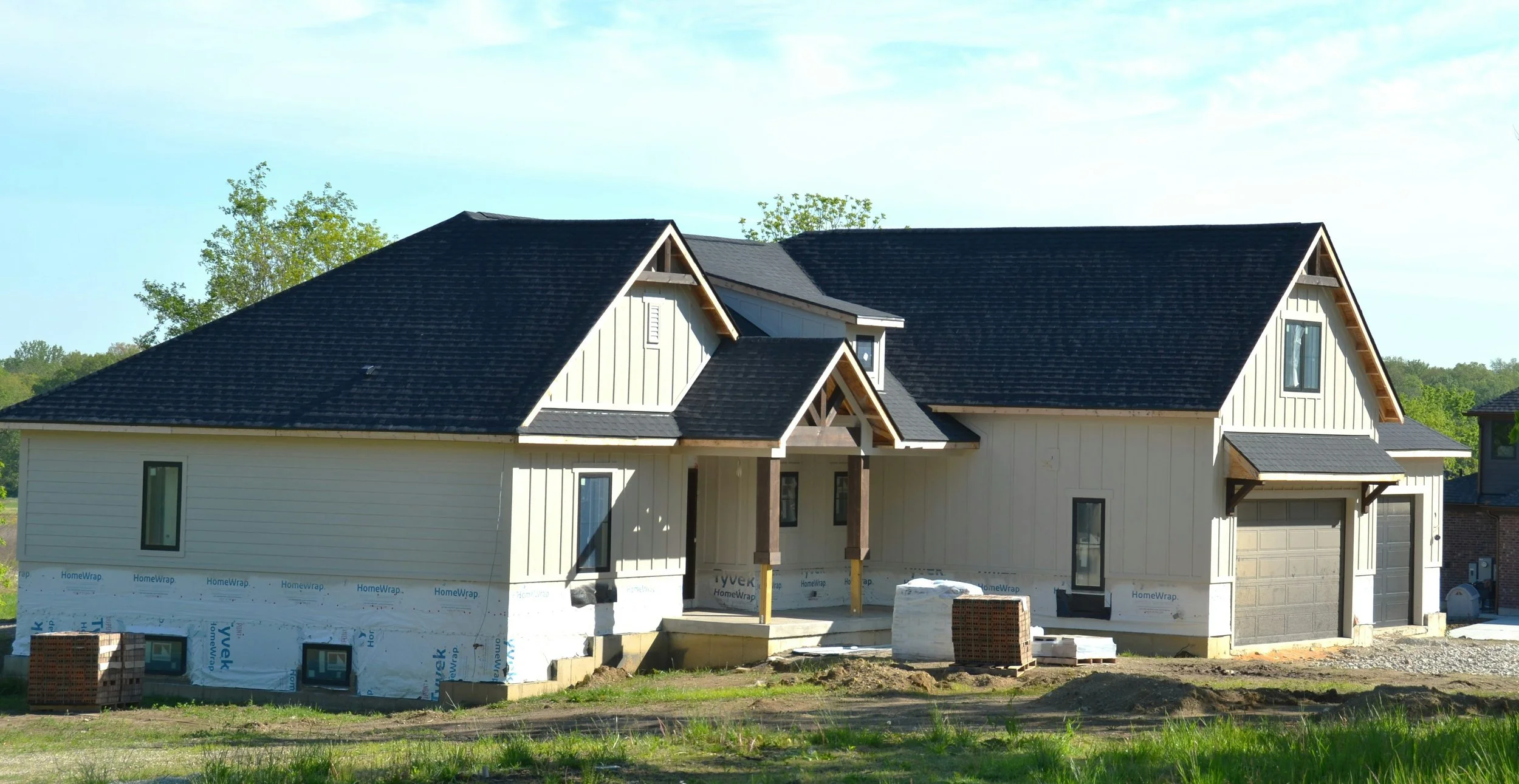 A house under construction with beige siding, black roof, and a garage, surrounded by construction materials and with trees and sky in the background.