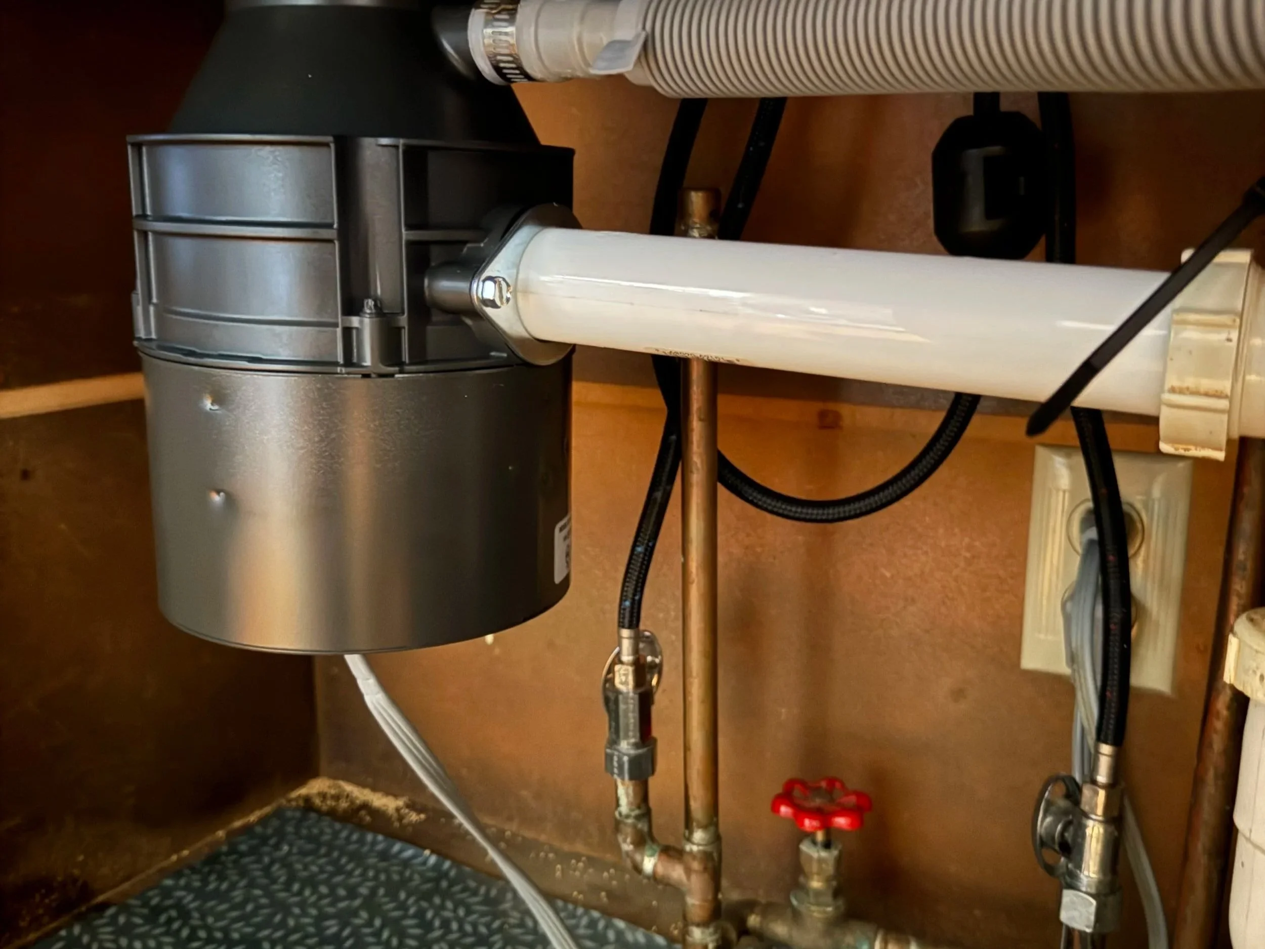 Underneath a sink with exposed plumbing, including pipes and a shut-off valve, located inside a cabinet with a wooden back wall.