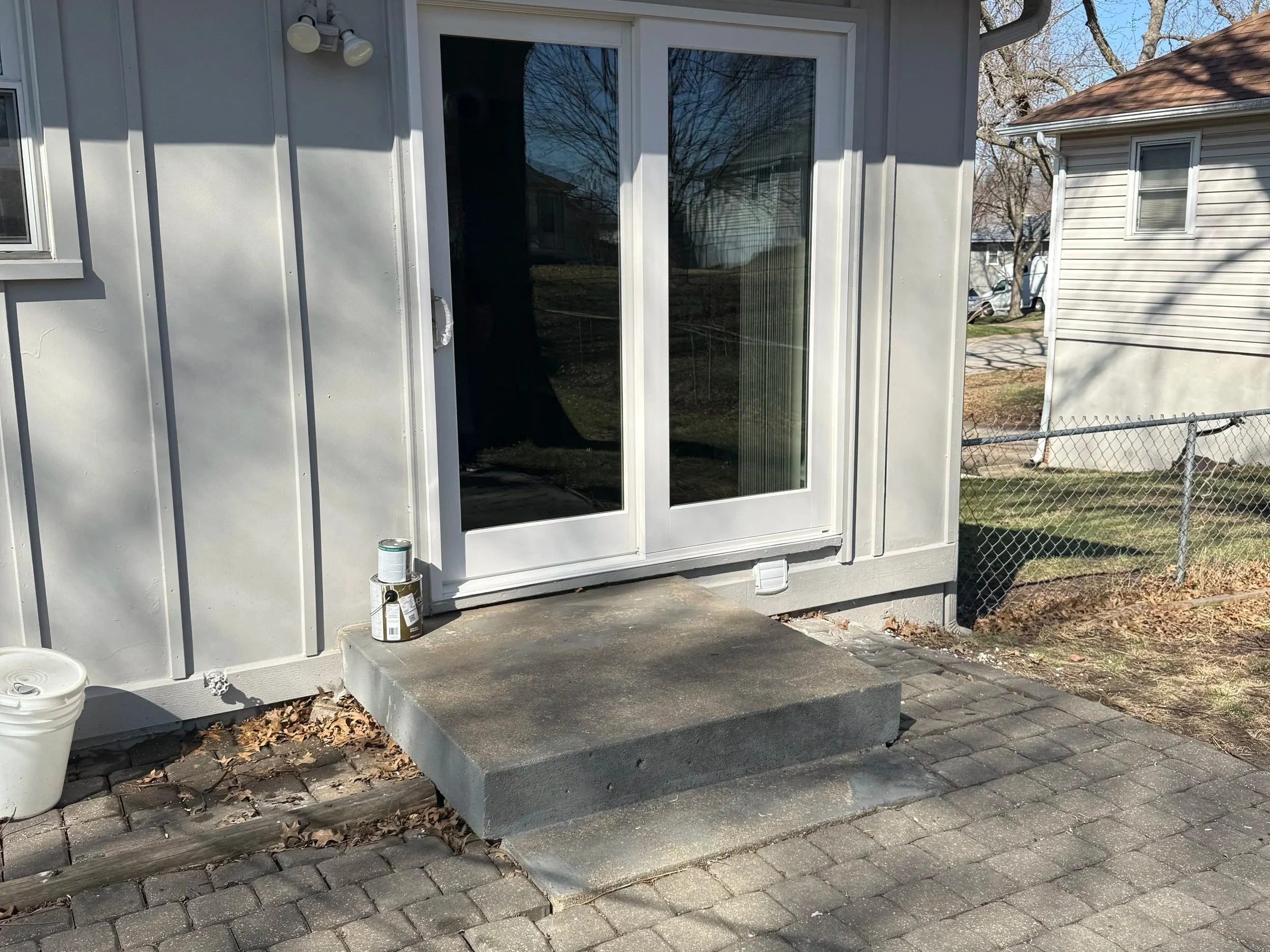 Backyard patio with new gray concrete steps leading to a sliding glass door, with a chain-link fence and neighboring houses in the background. Tools and paint cans are nearby.