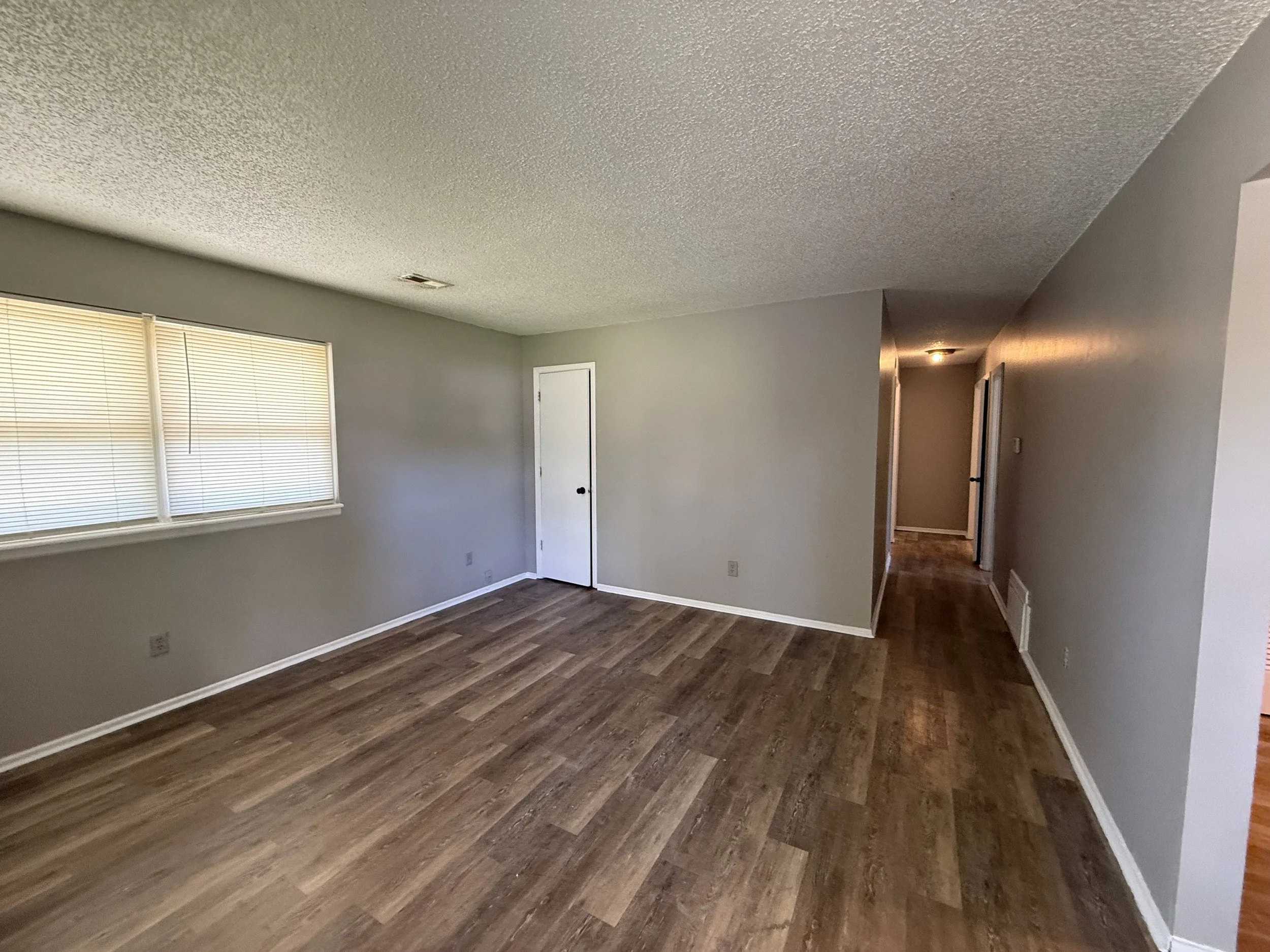 An empty living room with hardwood floors, light-colored walls, a window with blinds, and a white door.