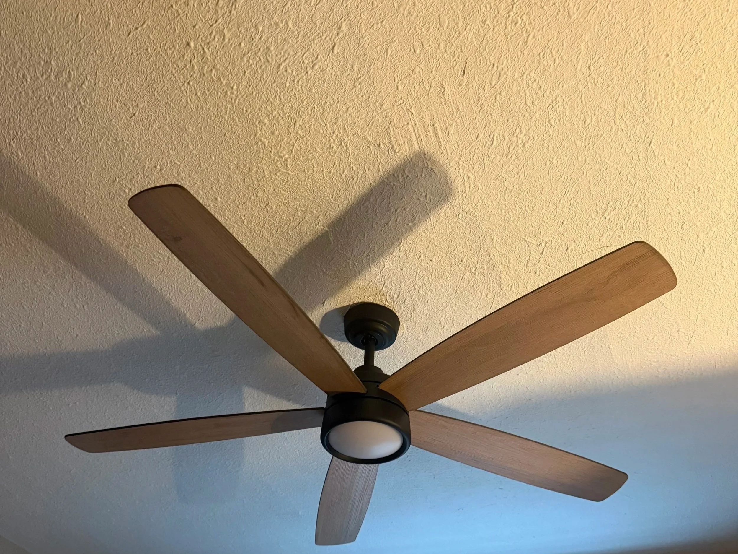 A ceiling fan with wooden blades and a light fixture in the center, mounted on a textured ceiling.
