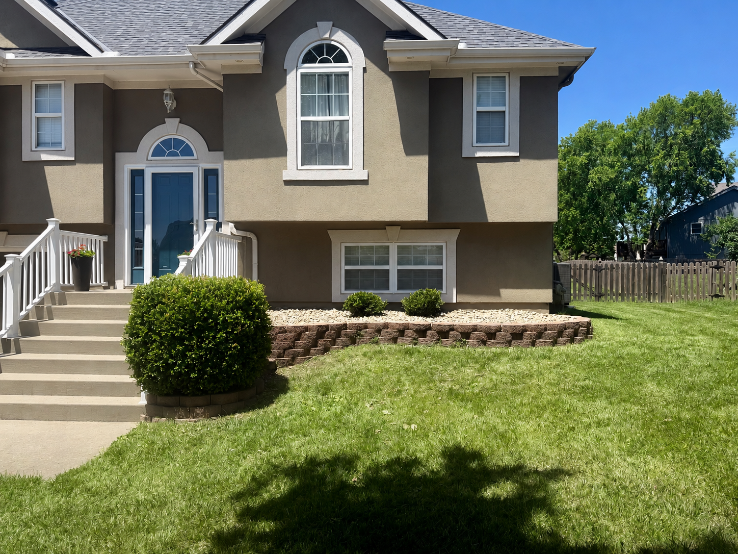 Front view of a beige two-story house with a gray roof, white trim, and a front stairway leading to a blue door, surrounded by a well-maintained green lawn and bushes, with a clear blue sky and trees in the background.