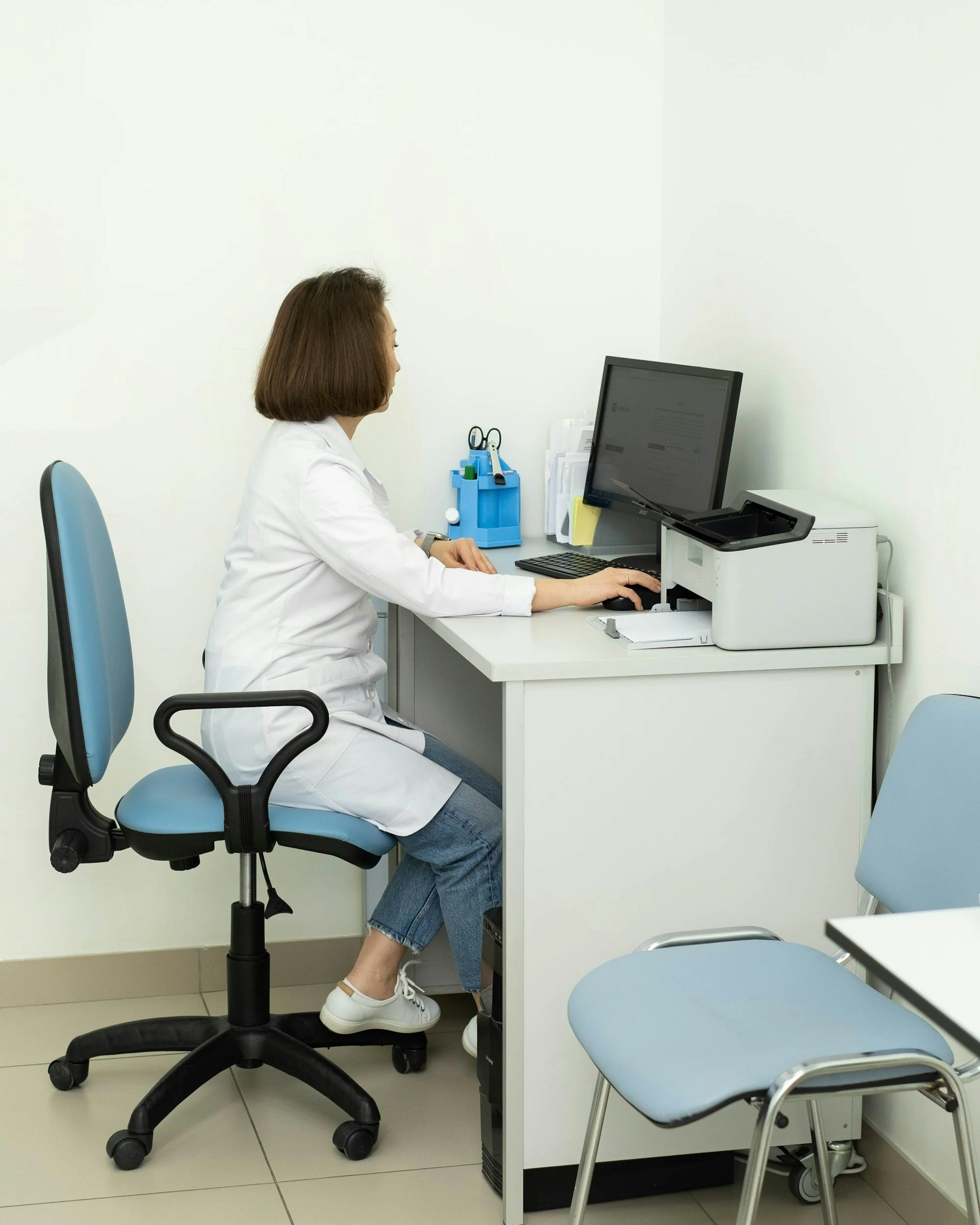 A woman in a white coat sitting at a desk in an office, working on a computer with a monitor, with a blue chair, printer, and various office supplies on the desk, against a plain white wall.