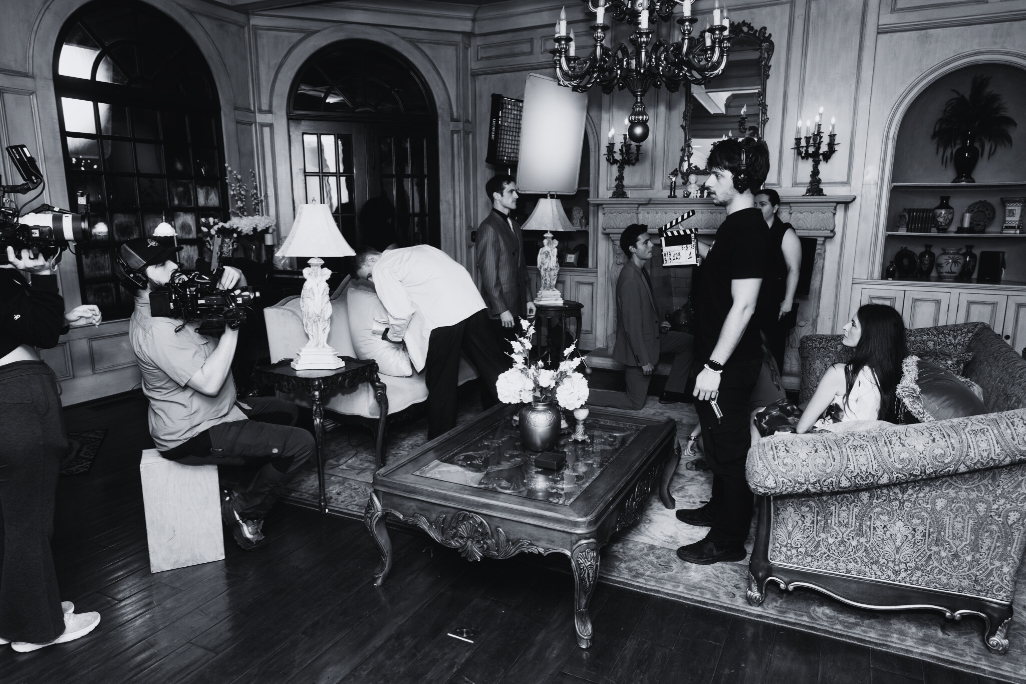 Filming crew setting up scene in a luxurious living room with vintage furniture, chandeliers, and decorative vases.