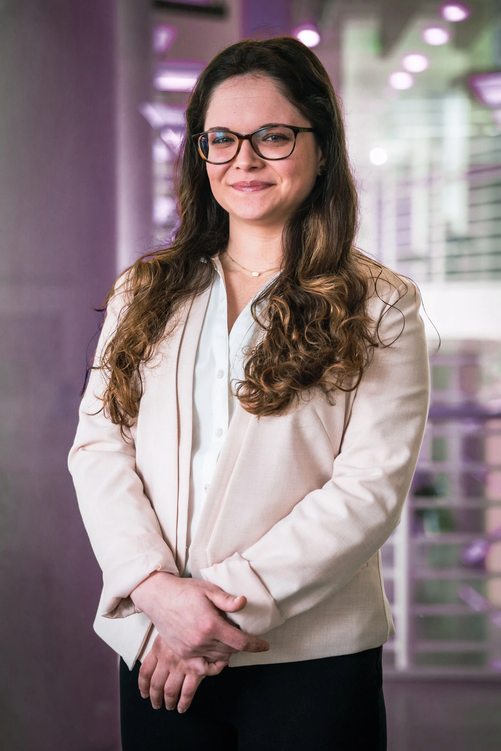 A woman with long wavy brown hair, wearing glasses, a white shirt, and a light beige blazer, standing with her hands clasped in front of her, smiling in an indoor setting with purple-lit decor and large windows in the background.