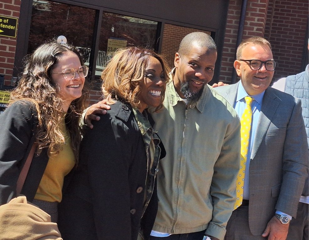 Four people standing close together outdoors, smiling. One woman has curly hair and glasses, another woman has shoulder-length hair, a man in a light jacket, and a man in a suit with glasses and a yellow tie.