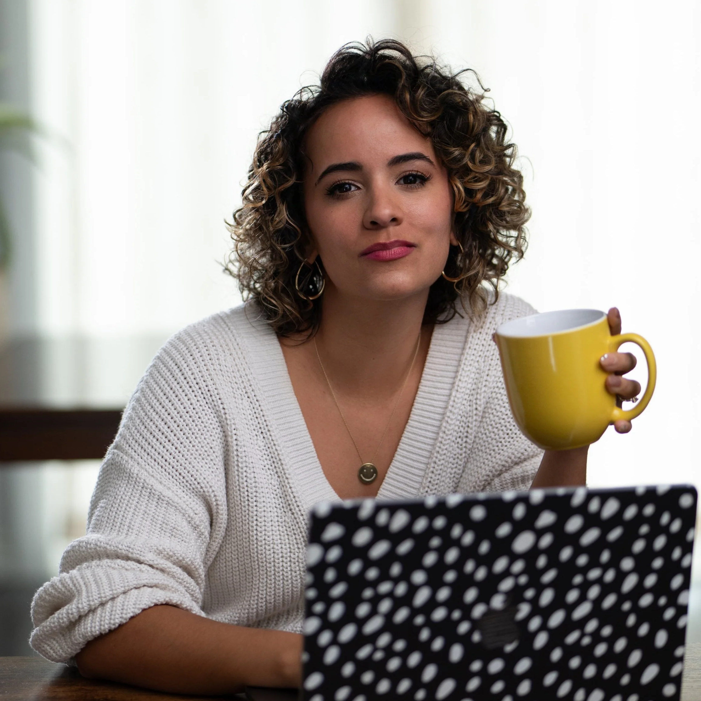 A woman with curly hair holding a yellow mug, sitting at a table with a polka dot laptop cover in a bright room.