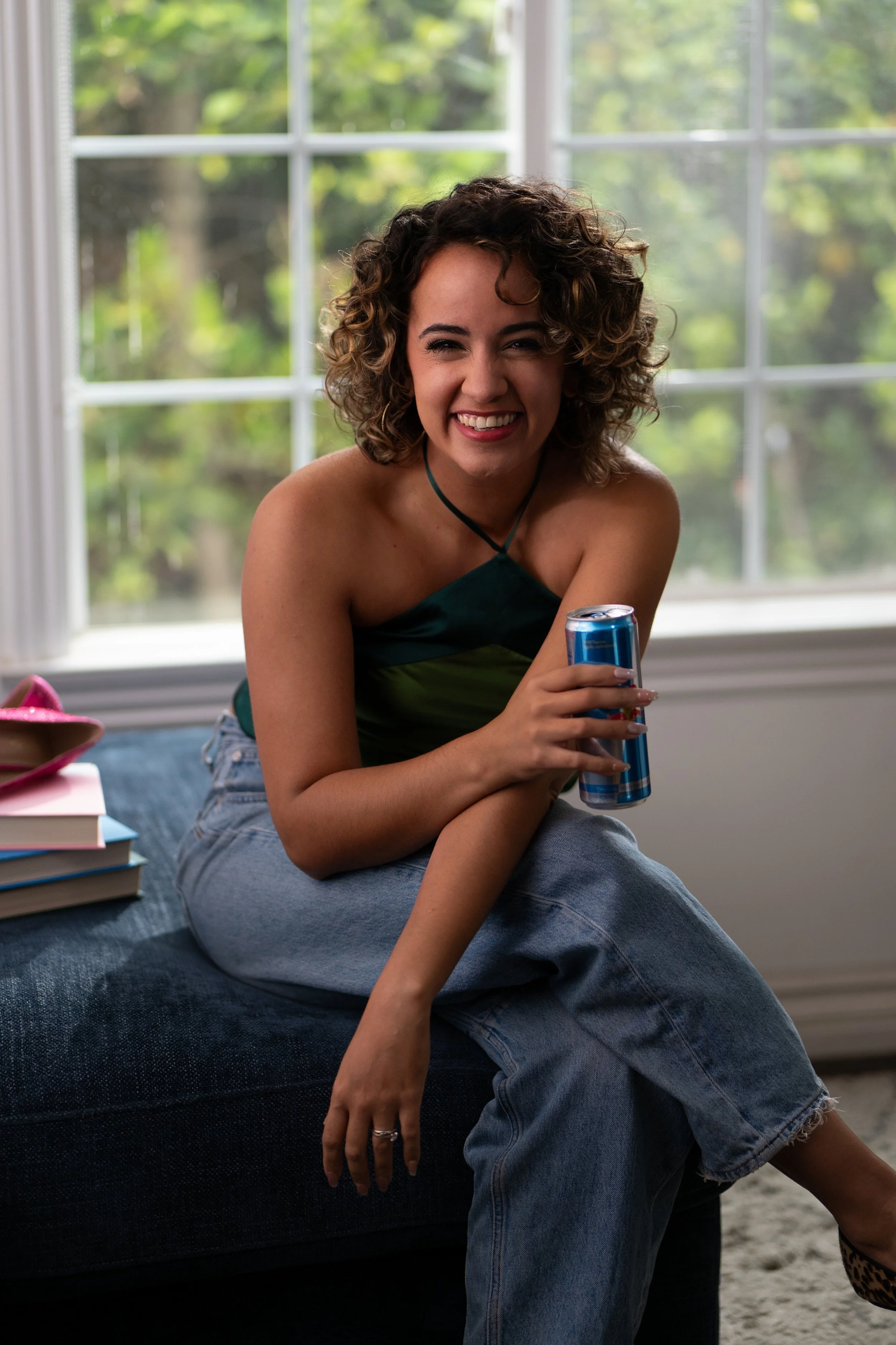 A woman with curly hair smiling and holding a soda can, sitting on a bed with a window and green trees outside in the background.