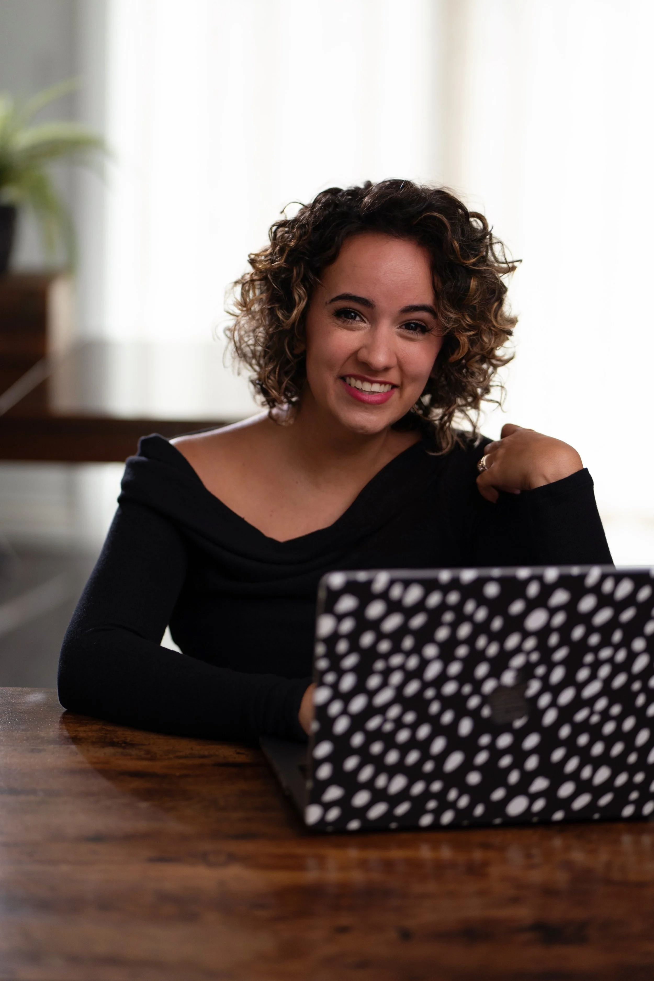 A woman with curly hair sitting at a wooden table using a laptop with a black and white polka dot cover. She is smiling and looking at the camera.
