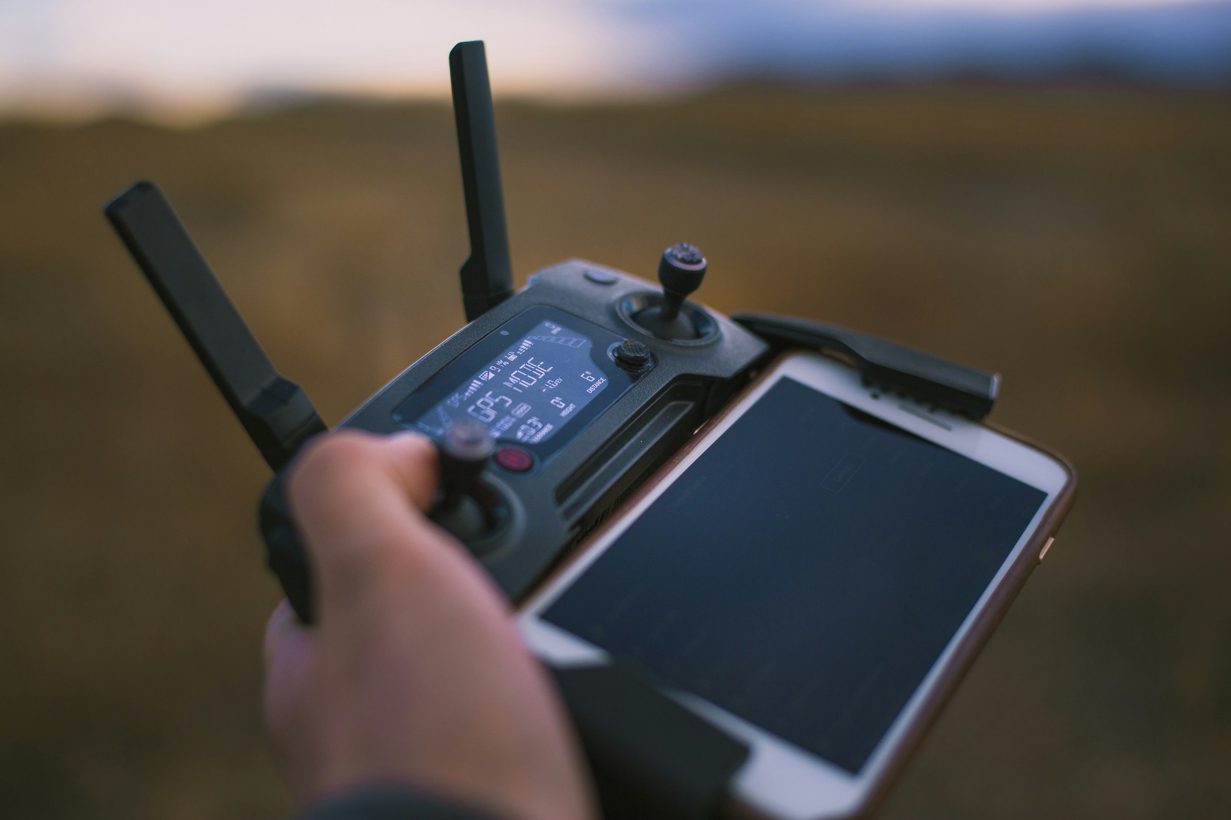 Close-up of a person's hand holding a drone remote controller with a smartphone attached, showing a landscape with a road and grass in the background.
