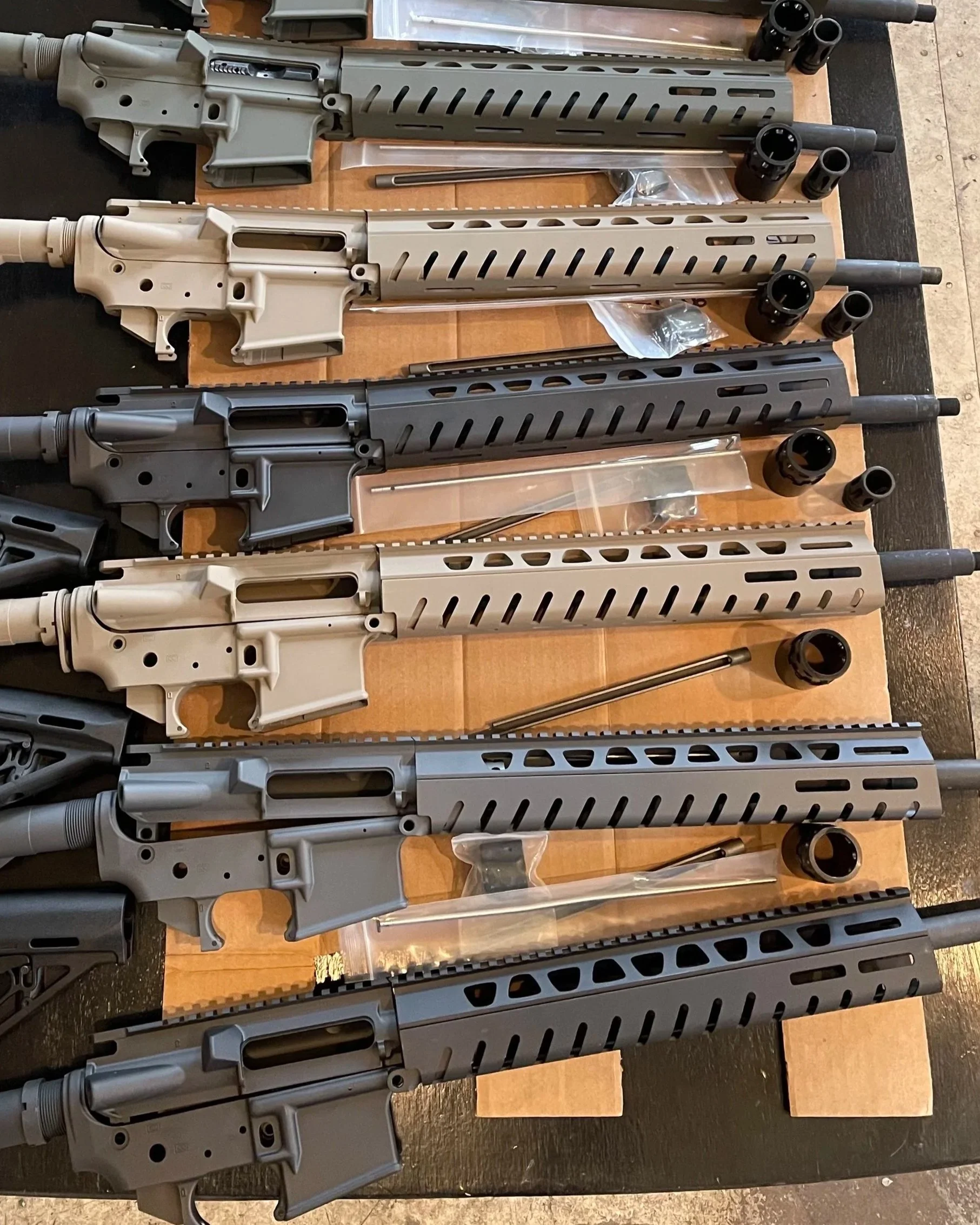 Multiple rifle upper receivers in different colors (tan, gray, black) with handguards, barrels, and various small parts laid out on a wooden table.