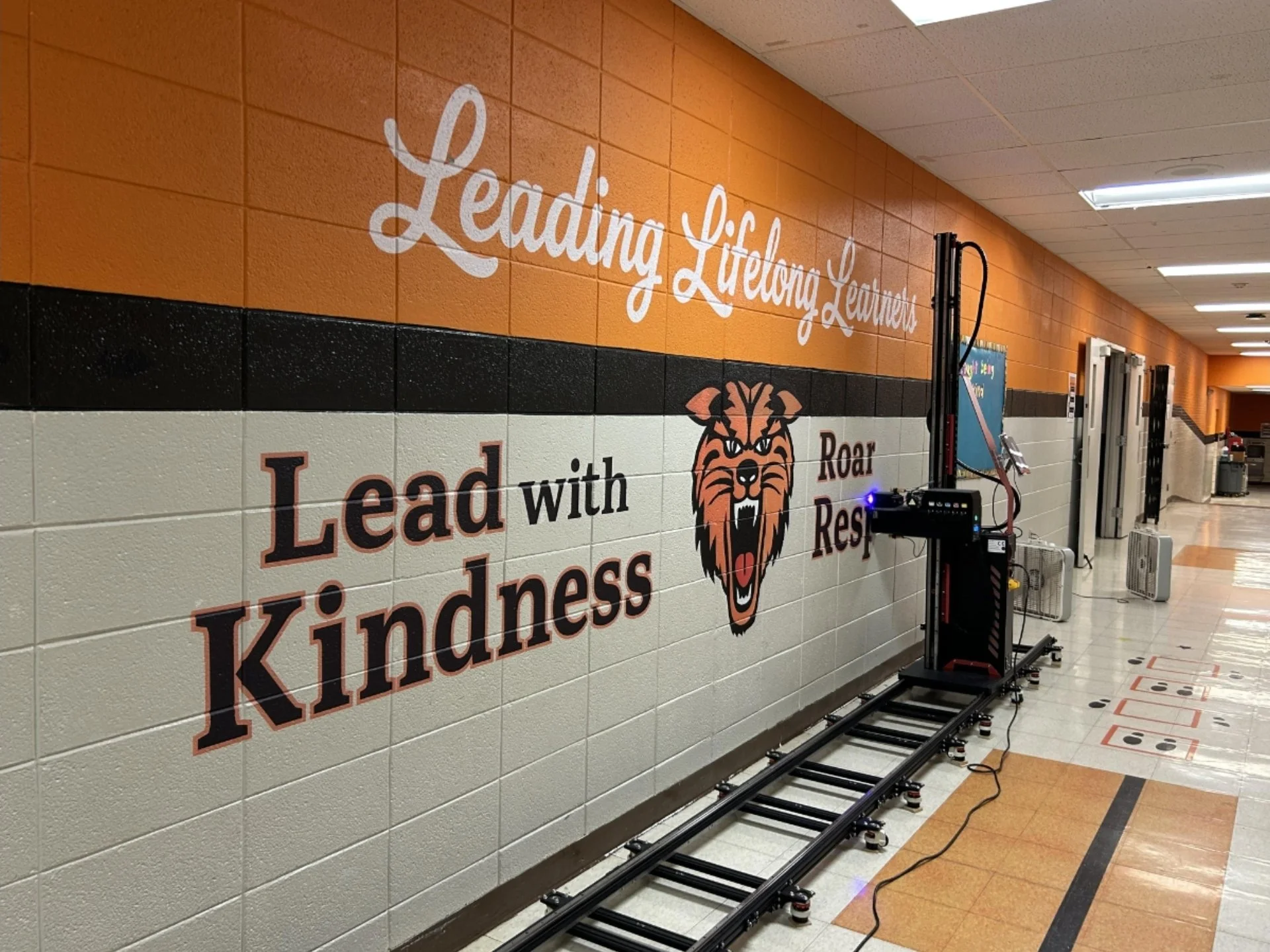 School hallway with orange and white tiled wall, large sign with motivational phrases, a robotic device on tracks, and lockers in the background.