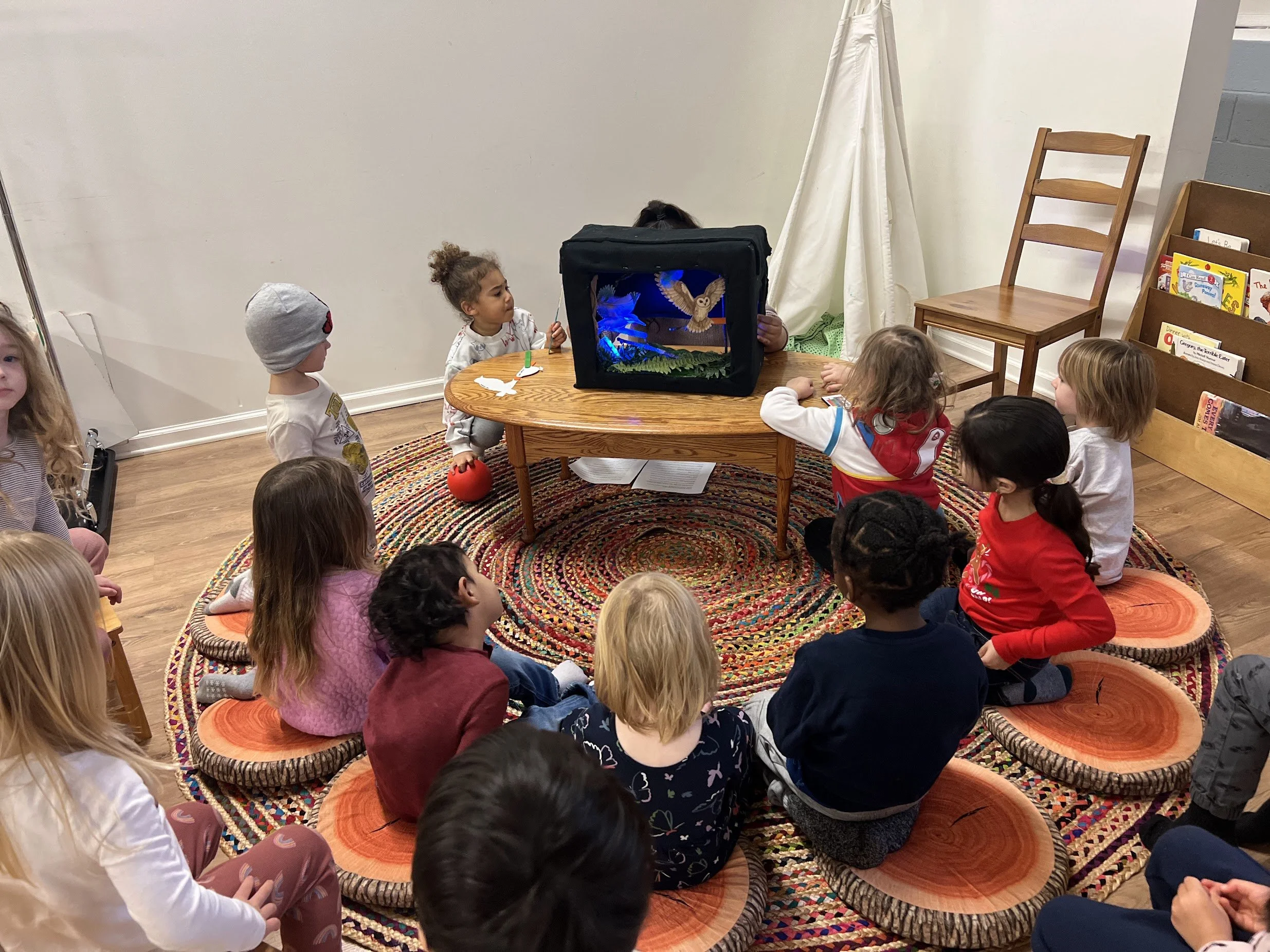children sitting on a rainbow circle rug watching a puppet show during art class
