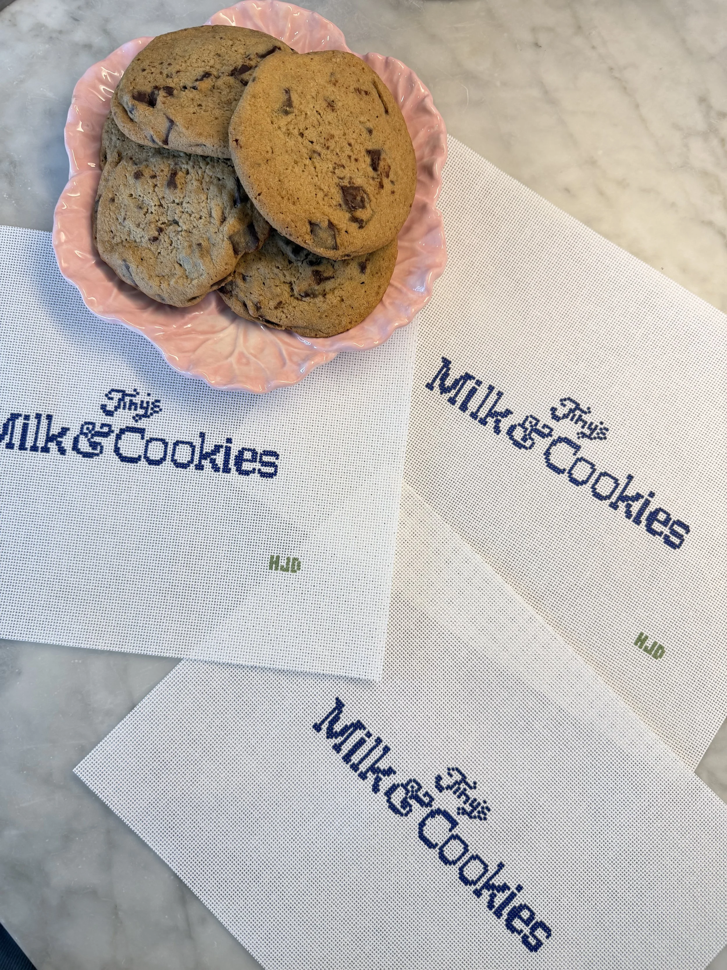A pink decorative plate with four chocolate chip cookies on a white and blue embroidered napkin that says 'Milk & Cookies'.