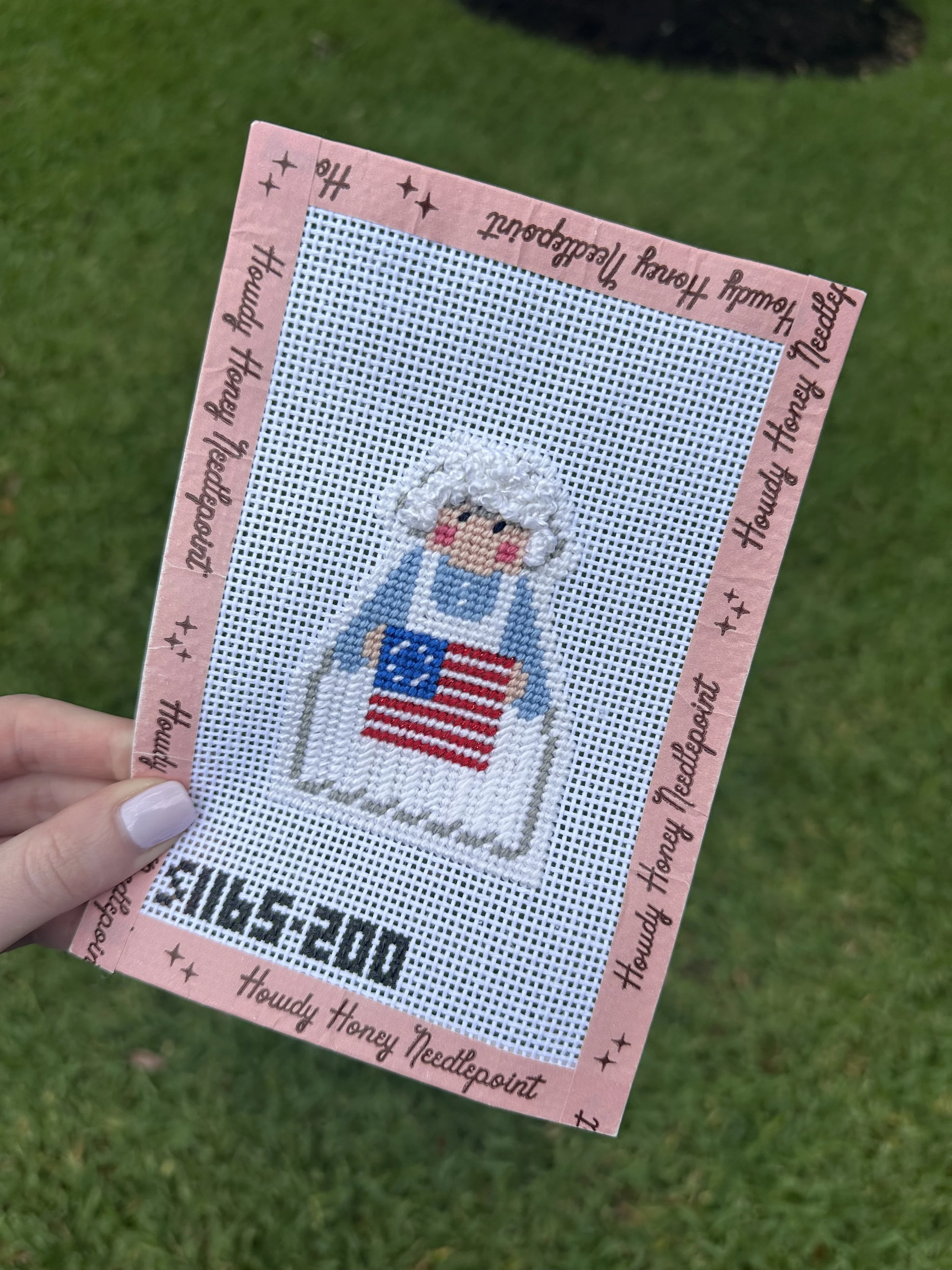 Cross-stitch embroidery of a girl wearing a blue dress with a red and white striped shirt, holding a small American flag, on a pink border with the words 'Houndy Honey Needlepoin' and star decorations.