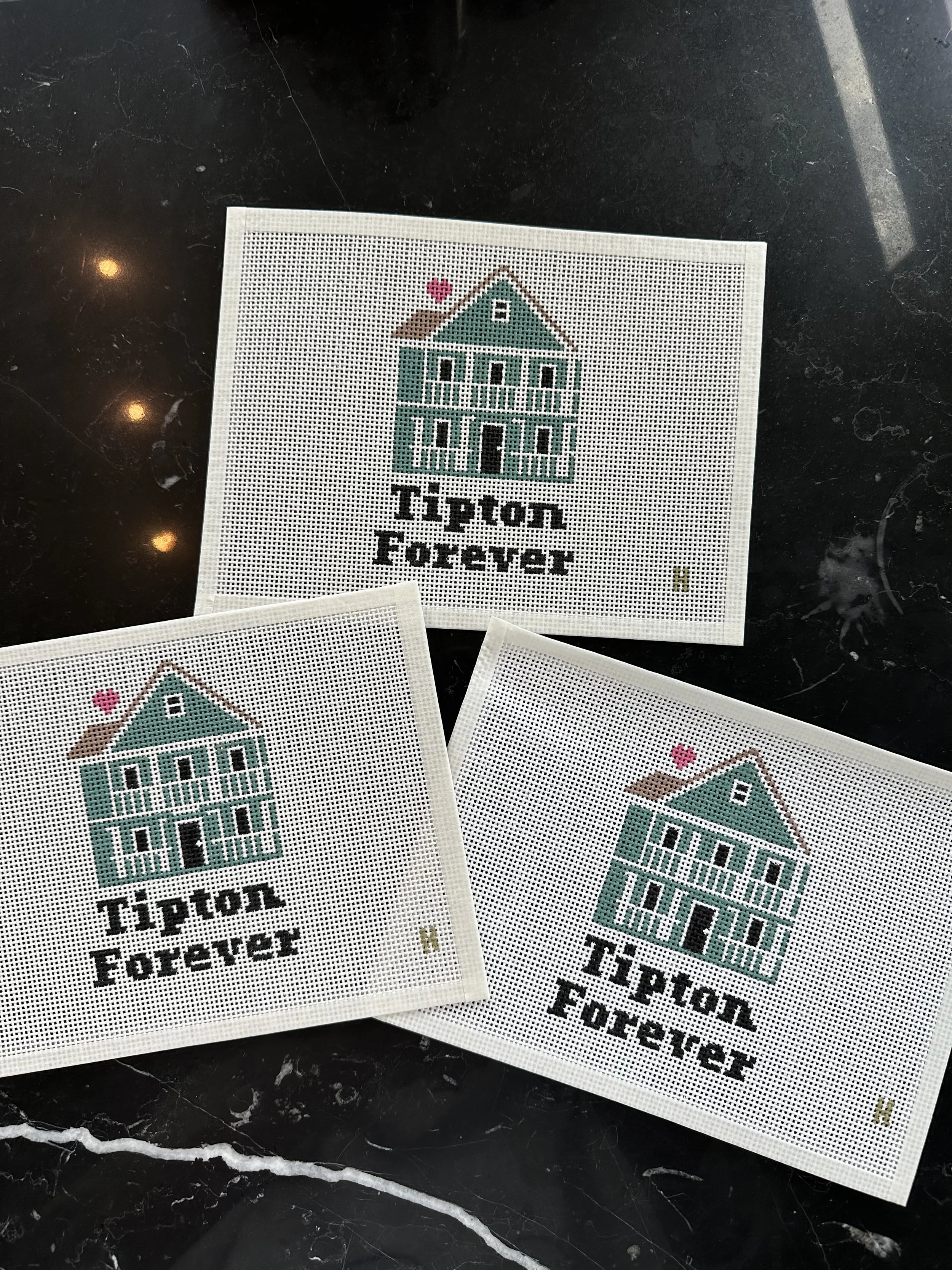 Cross-stitched fabric with a house and the phrase "Tipton Forever" printed underneath, laid on a black marble surface.