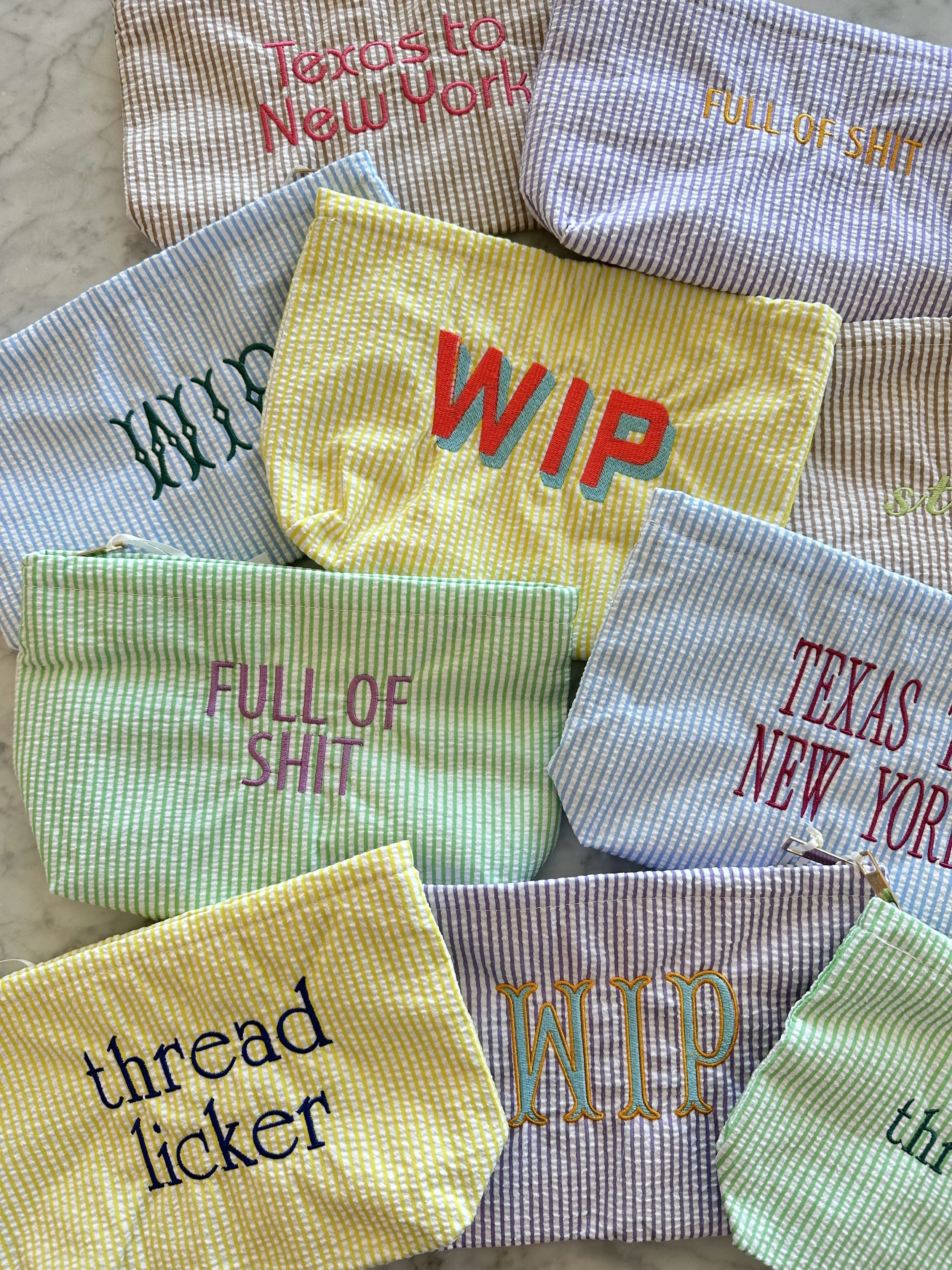 Multiple striped fabric pouches with embroidered text, including phrases like 'WIP,' 'full of shit,' 'Texas to New York,' and 'threaten' on various colored backgrounds.