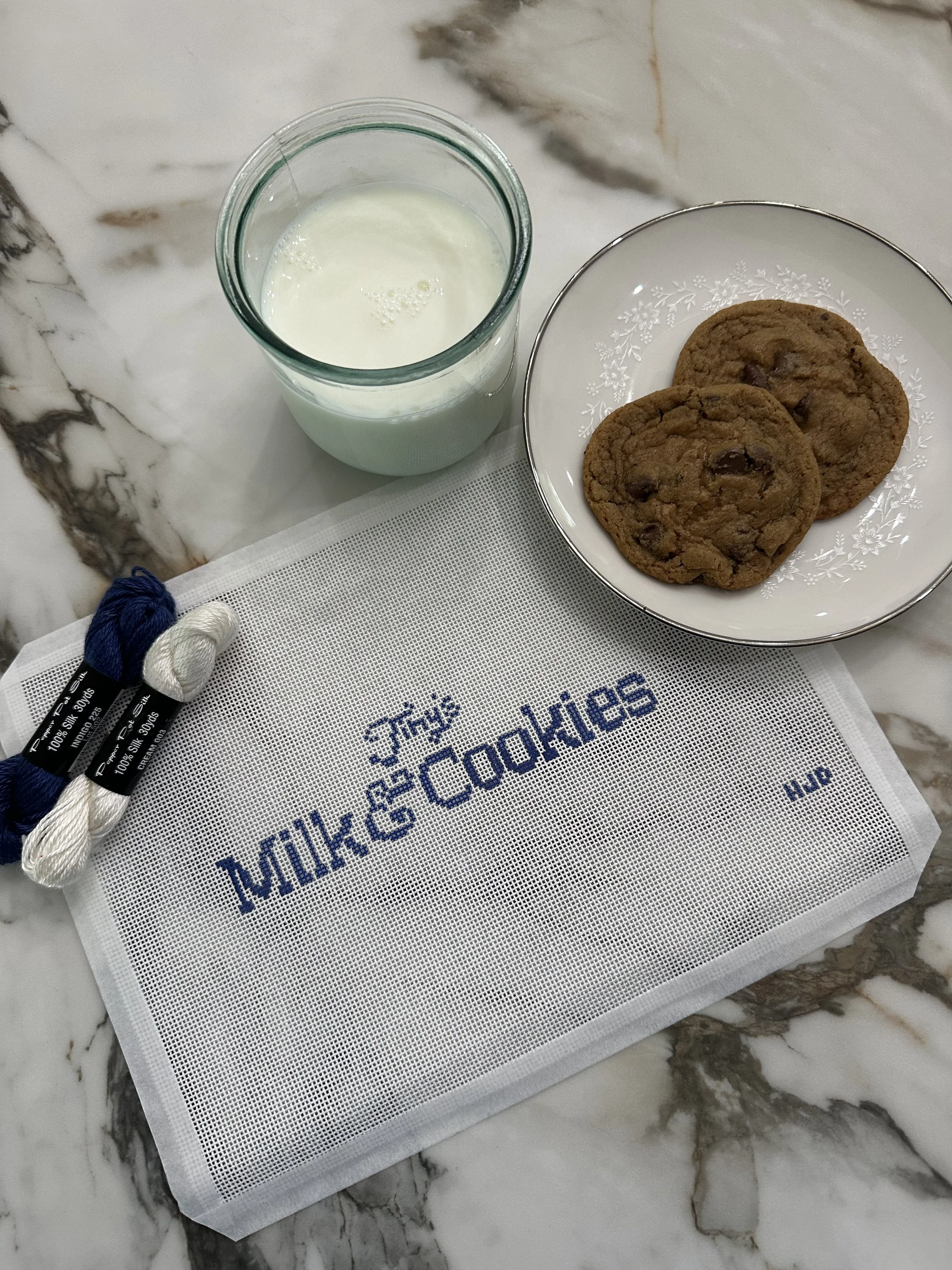 A glass of milk, a plate with two chocolate chip cookies, and a piece of white embroidery with blue text reading 'Milk & Cookies' on a marble surface.