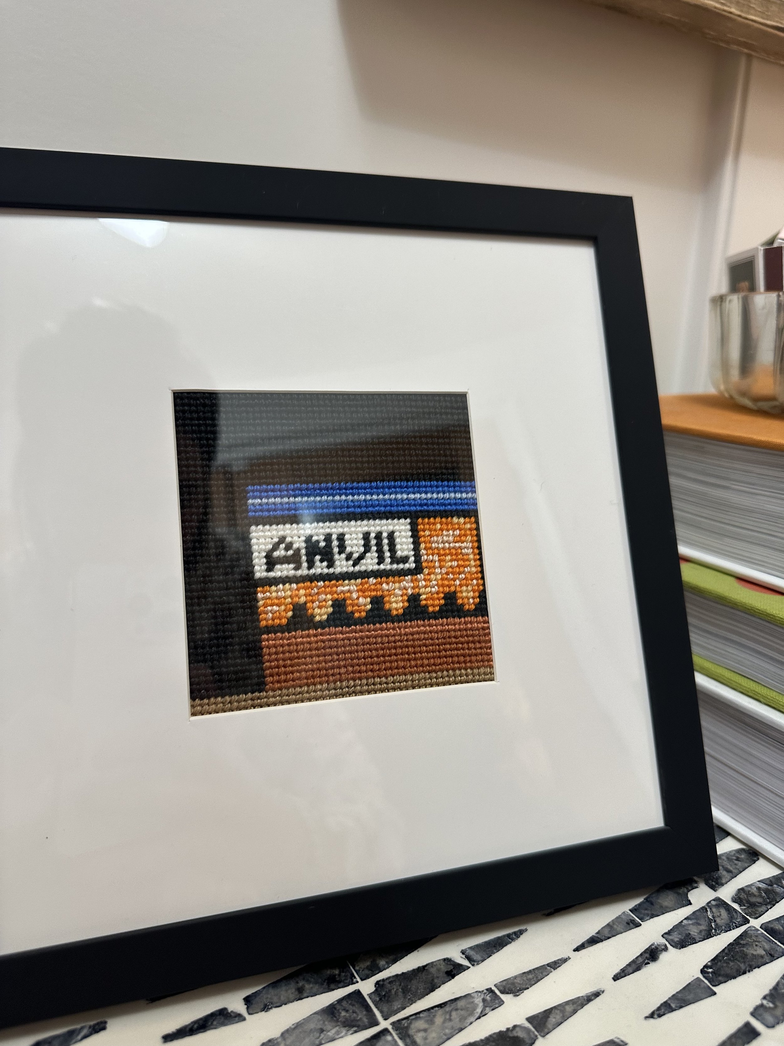 Close-up of a framed piece of craft or cross-stitch art with the text 'TEXAS' visible, resting on a white surface with a patterned black-and-white tile floor in the background.