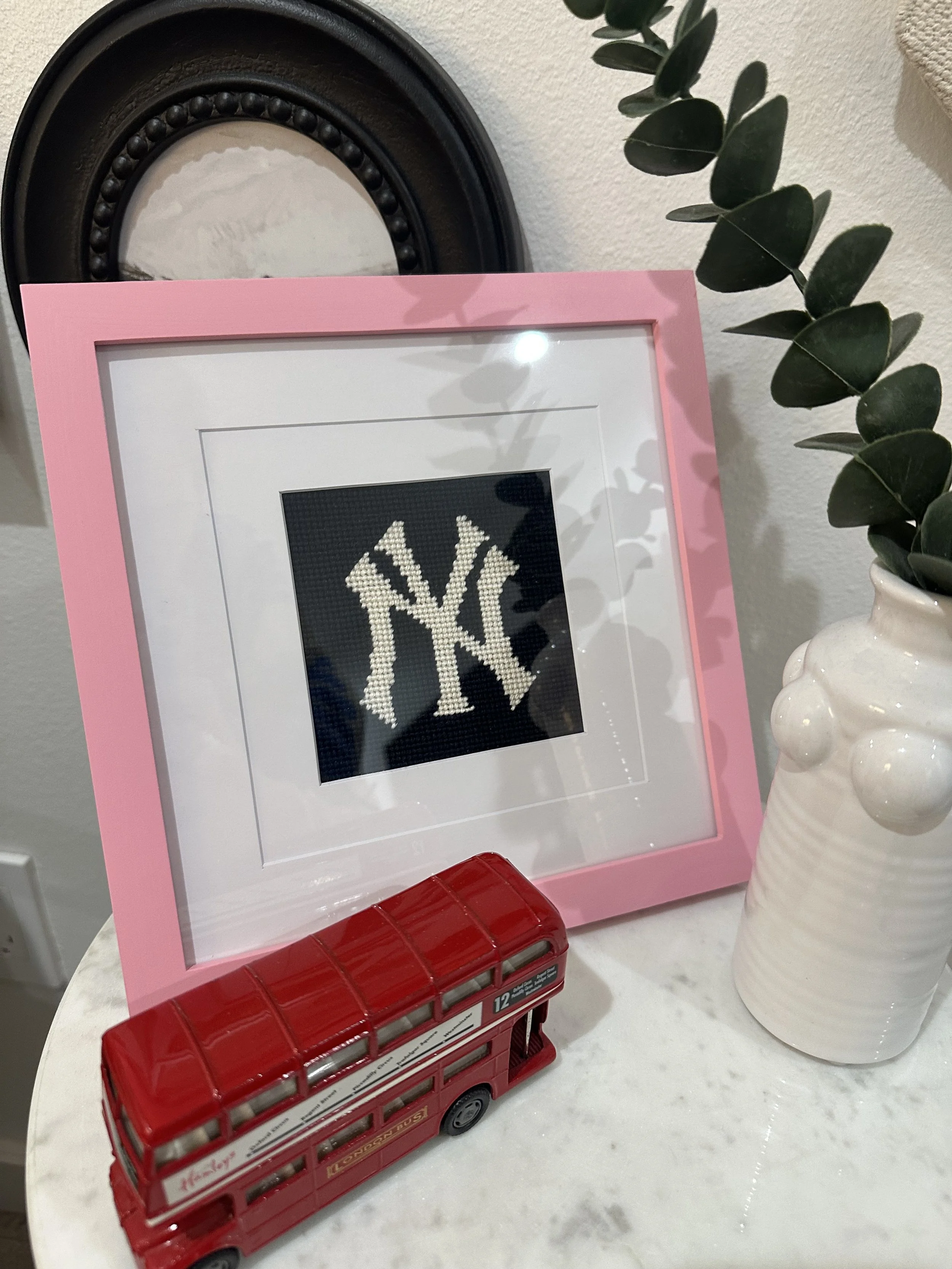 Decorative display on a white marble surface with a pink framed picture, a miniature red double-decker bus toy, and a white textured vase with green foliage.
