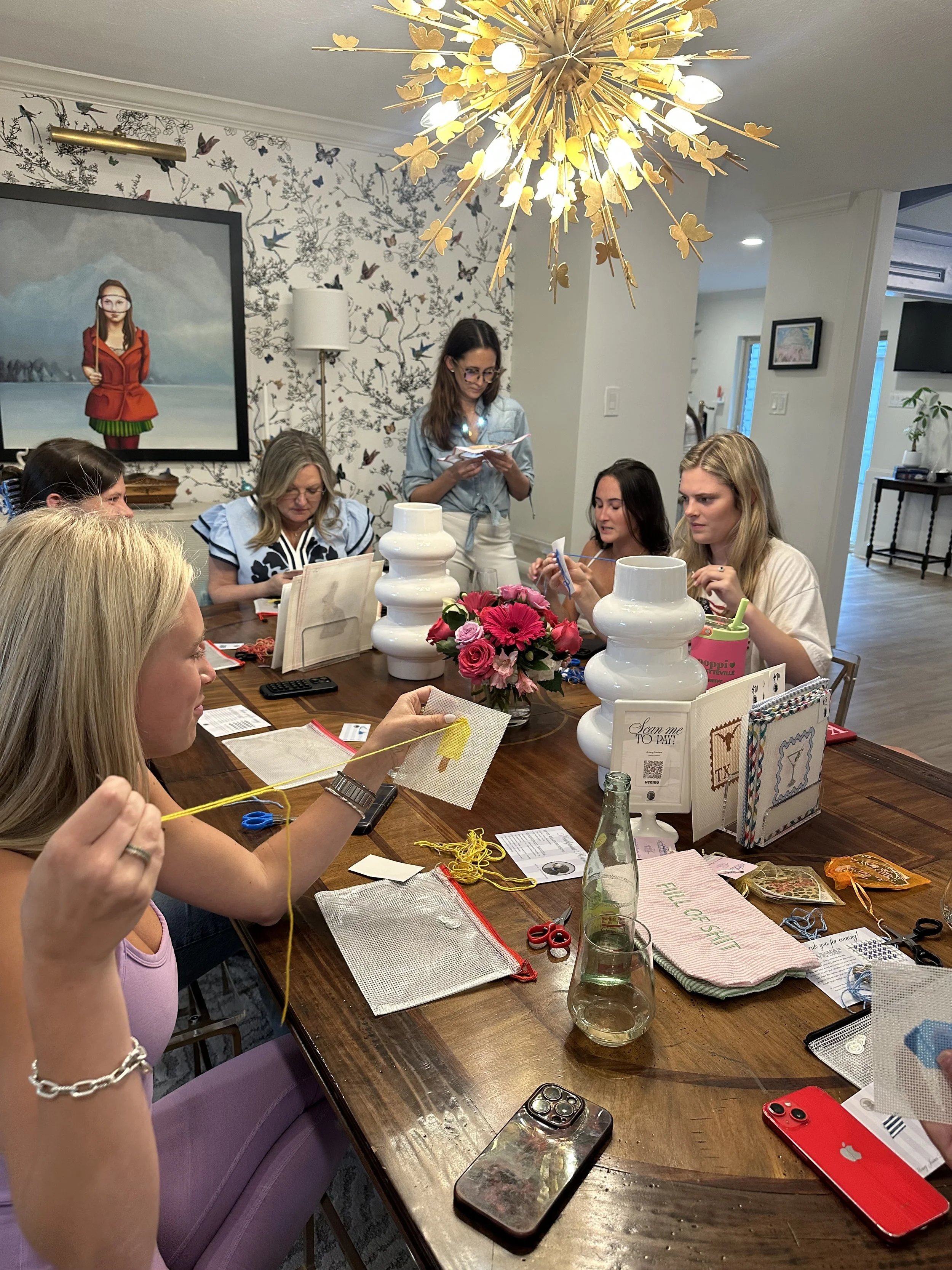 A group of women gathered around a wooden table, engaged in crafting or needlework activities. The table has various sewing supplies, notebooks, and a pink flower arrangement in the center. The room is decorated with butterfly-themed wallpaper and a 