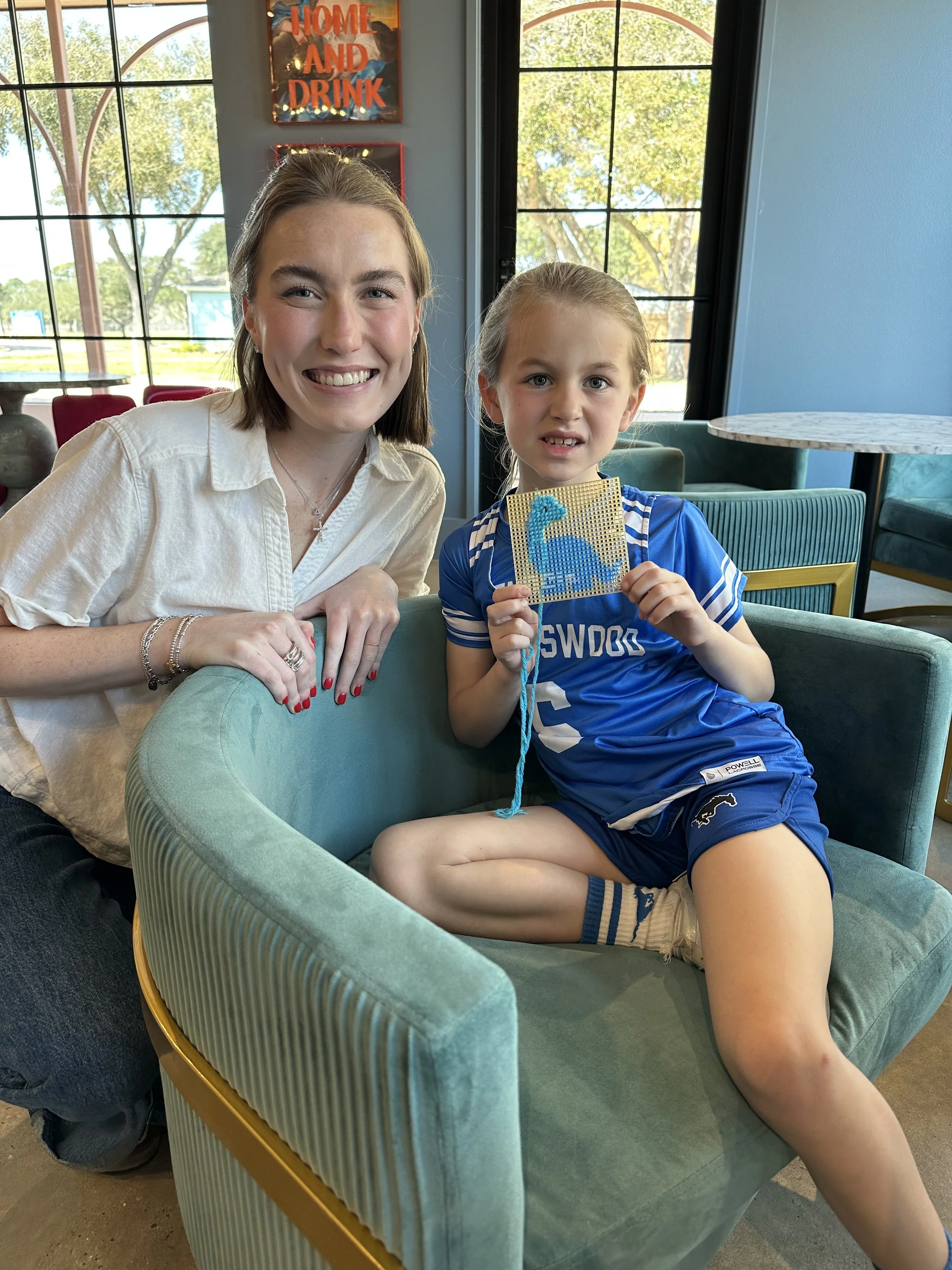A woman smiling sitting next to a young girl holding a blue beaded art piece in a cafe or restaurant with large windows showing outdoor trees.