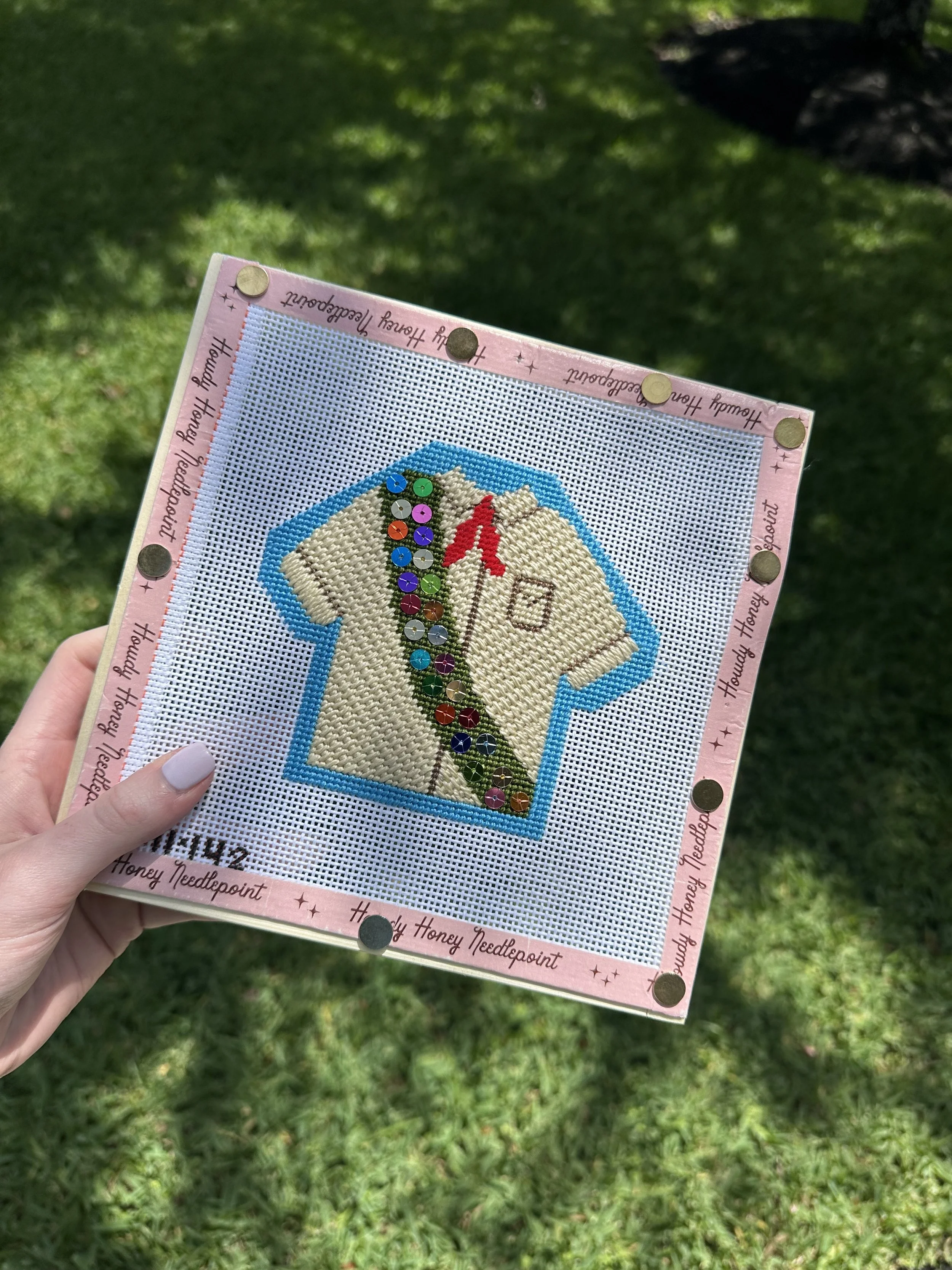 A hand holding a piece of cross-stitch embroidery on white fabric, featuring a beige puppy with a red bow. The embroidery is framed by a pink border with black text that reads 'Honey Honey Needlepoint' and decorative stars, with metallic circular emb