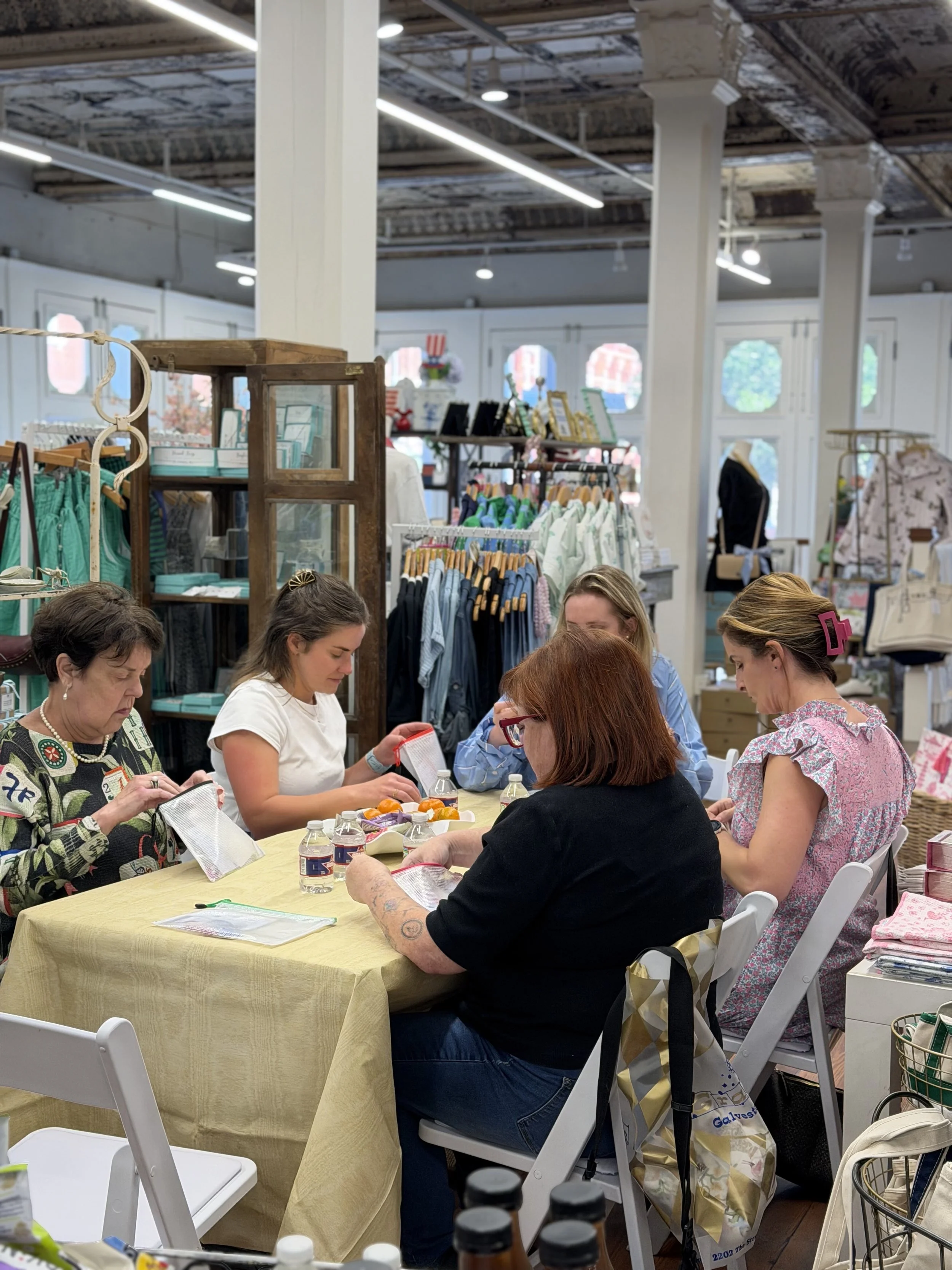People sitting around a table in a retail store, engaged in a craft or sewing activity, with clothing and accessories displayed in the background.