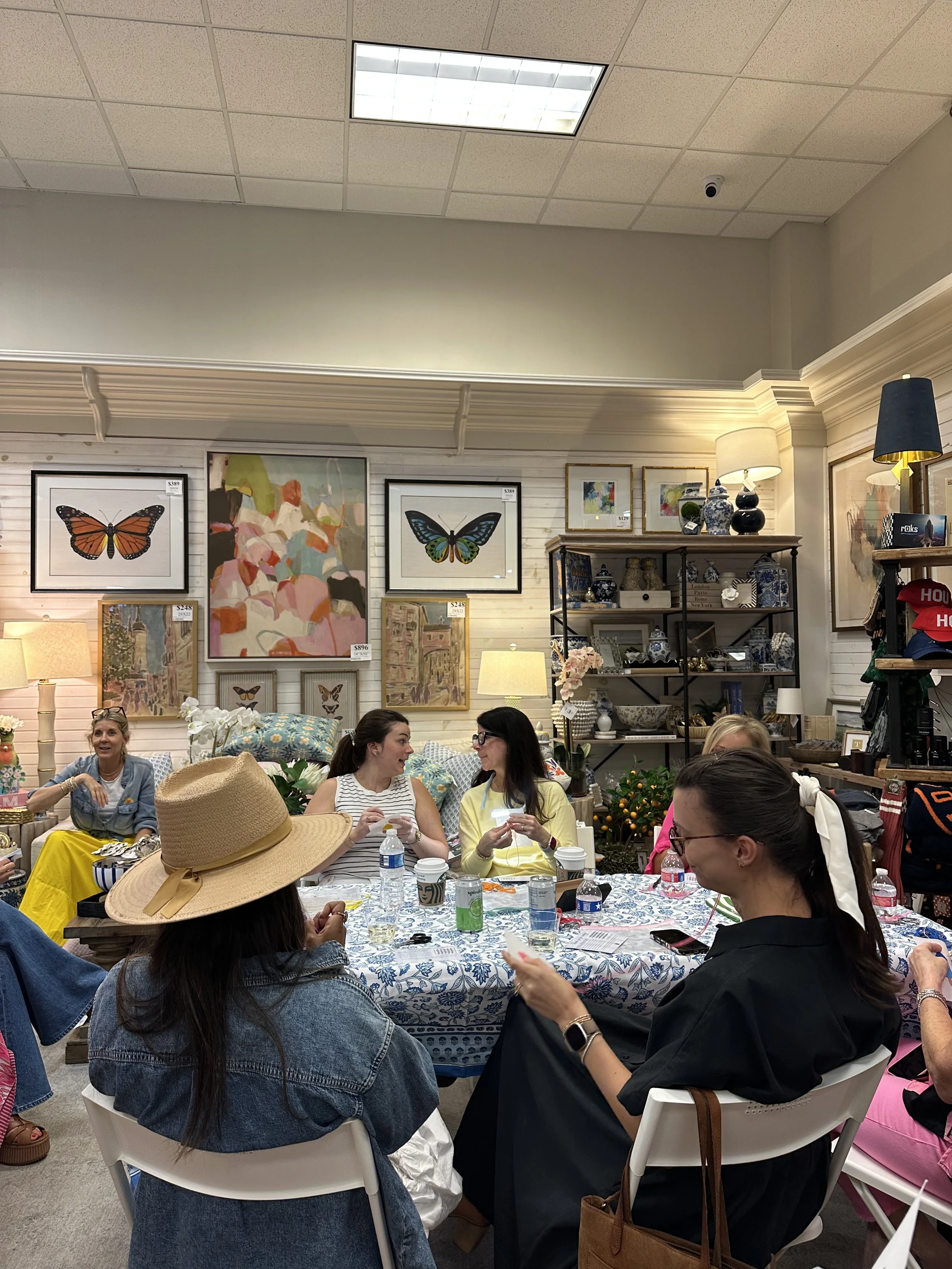 Group of women sitting around a table inside a store, engaged in conversation, with artwork and decorative items displayed on the walls and shelves behind them.