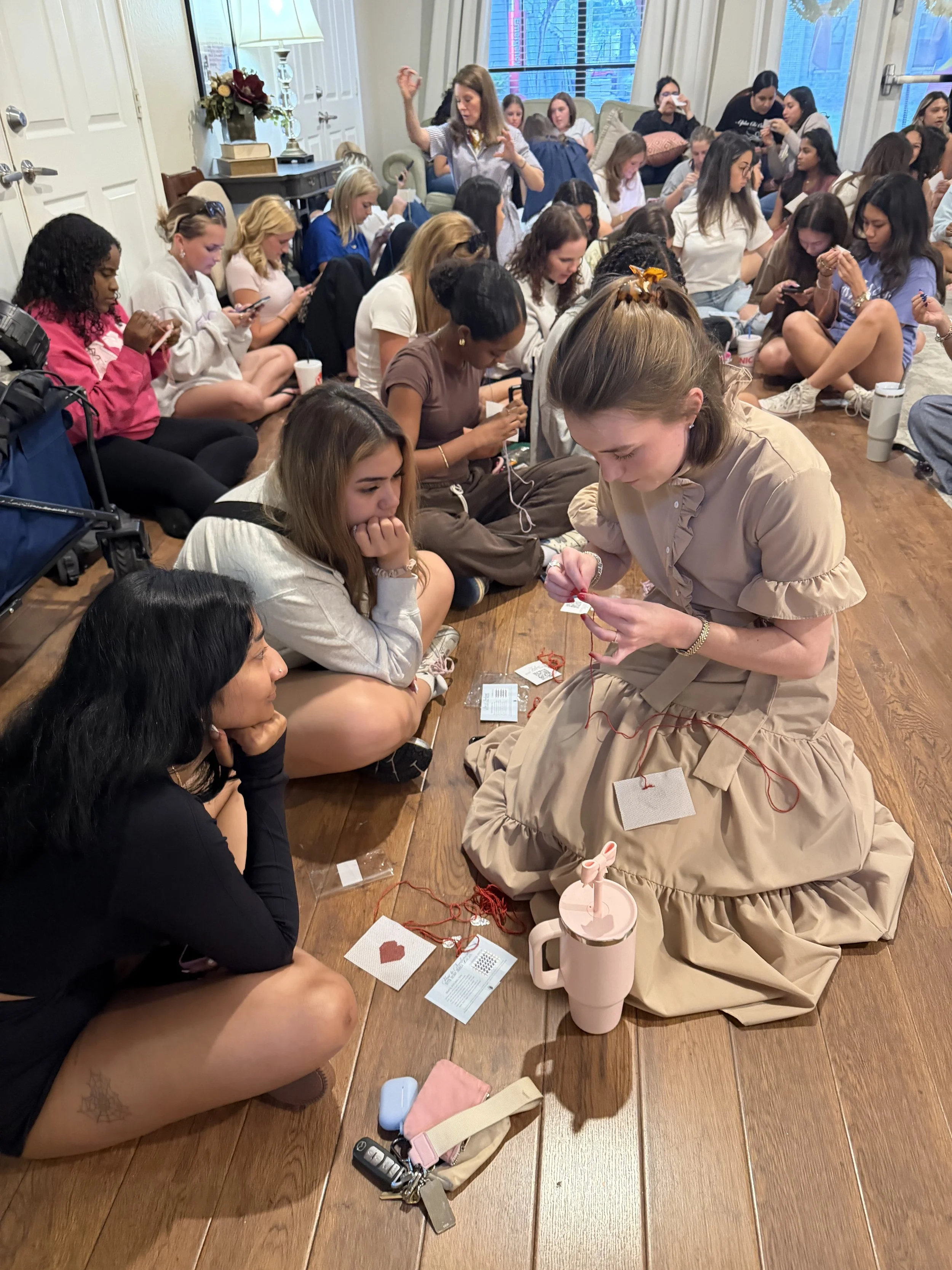 A group of women sitting on the floor in a room, many focused on their phones, with some engaged in crafting activities involving yarn and small craft supplies.