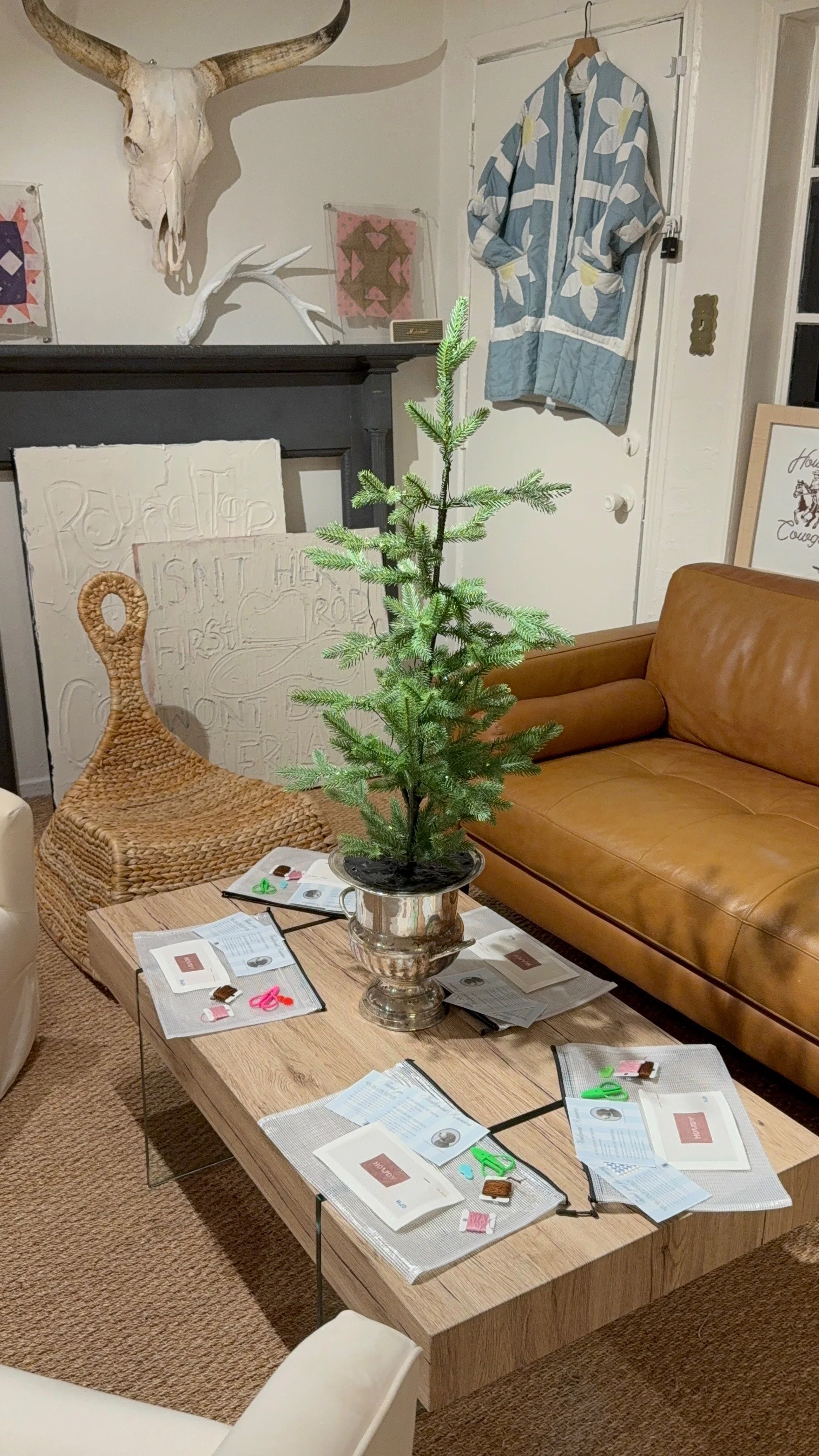 A small, young Christmas tree in a silver pot sits on a wooden coffee table, surrounded by scattered papers, scissors, and other small items. In the background, there's a tan leather sofa, a wicker chair, and a fireplace with a mounted cow skull and 