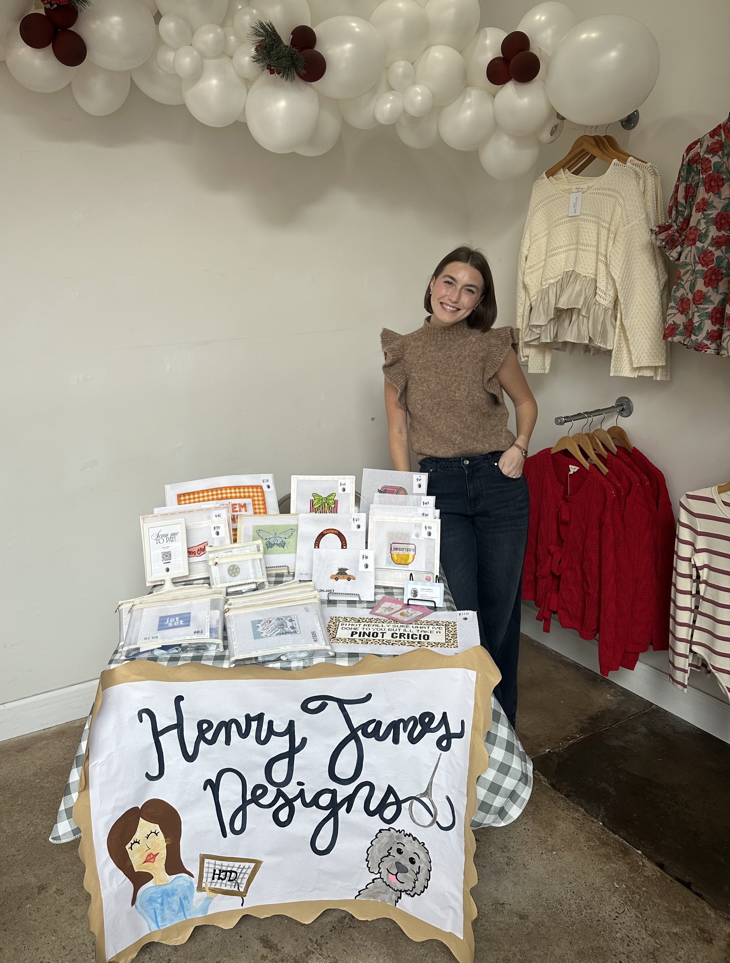 A young woman stands behind a table at a craft fair, smiling. The table displays embroidered fabric items such as napkins and accessories. Behind her, decorative clothes hang on the wall, including a yellow knit sweater and a floral-patterned dress. 