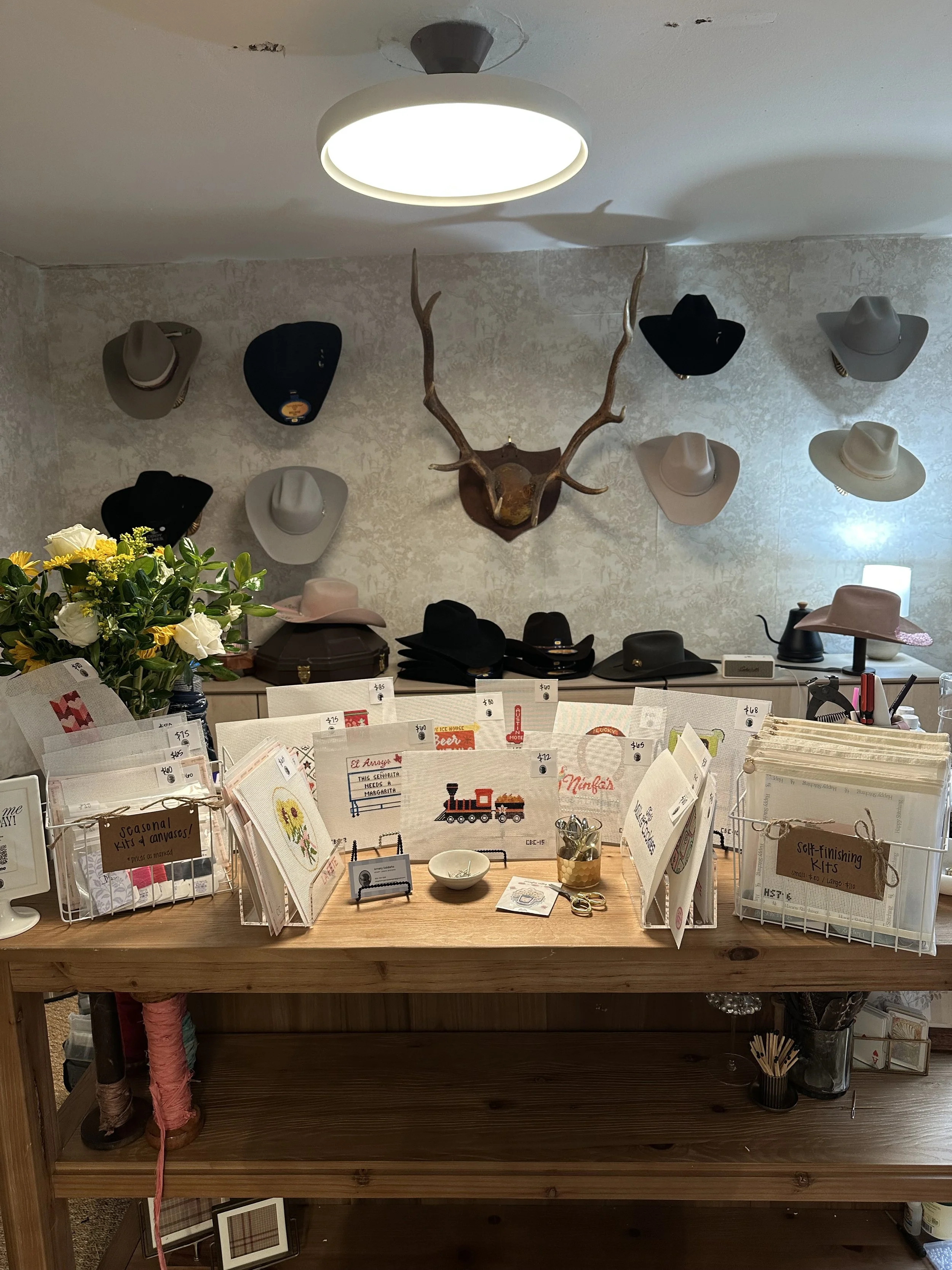 A display table with cross-stitch kits and embroidery supplies in front of a wall with mounted hats and a deer head with antlers. There is a flower arrangement on the left.