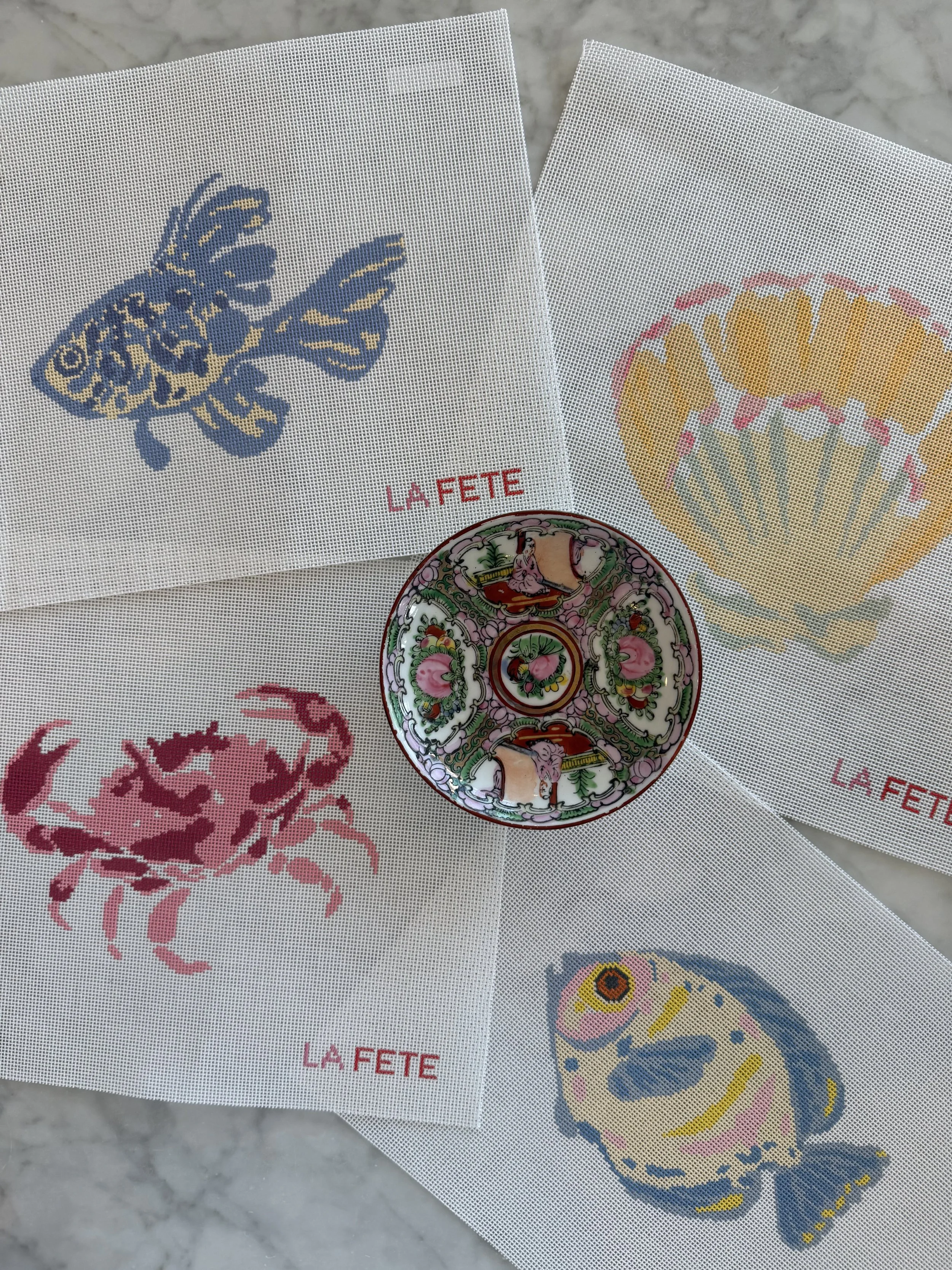 Several white cloth napkins with cross-stitch embroidery of colorful butterflies, crabs, and stylized floral designs, with文字 La Fete printed on them, laid out on a marble surface.