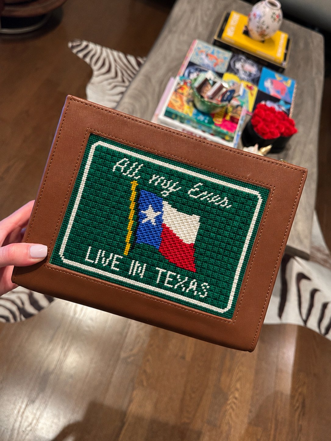 A stitched patch on a brown handbag displaying the Texas flag, the words "All Way Ends Love in Texas," and a yellow and blue vertical line, held over a table with magazines, a small vase, and a black container of red roses.