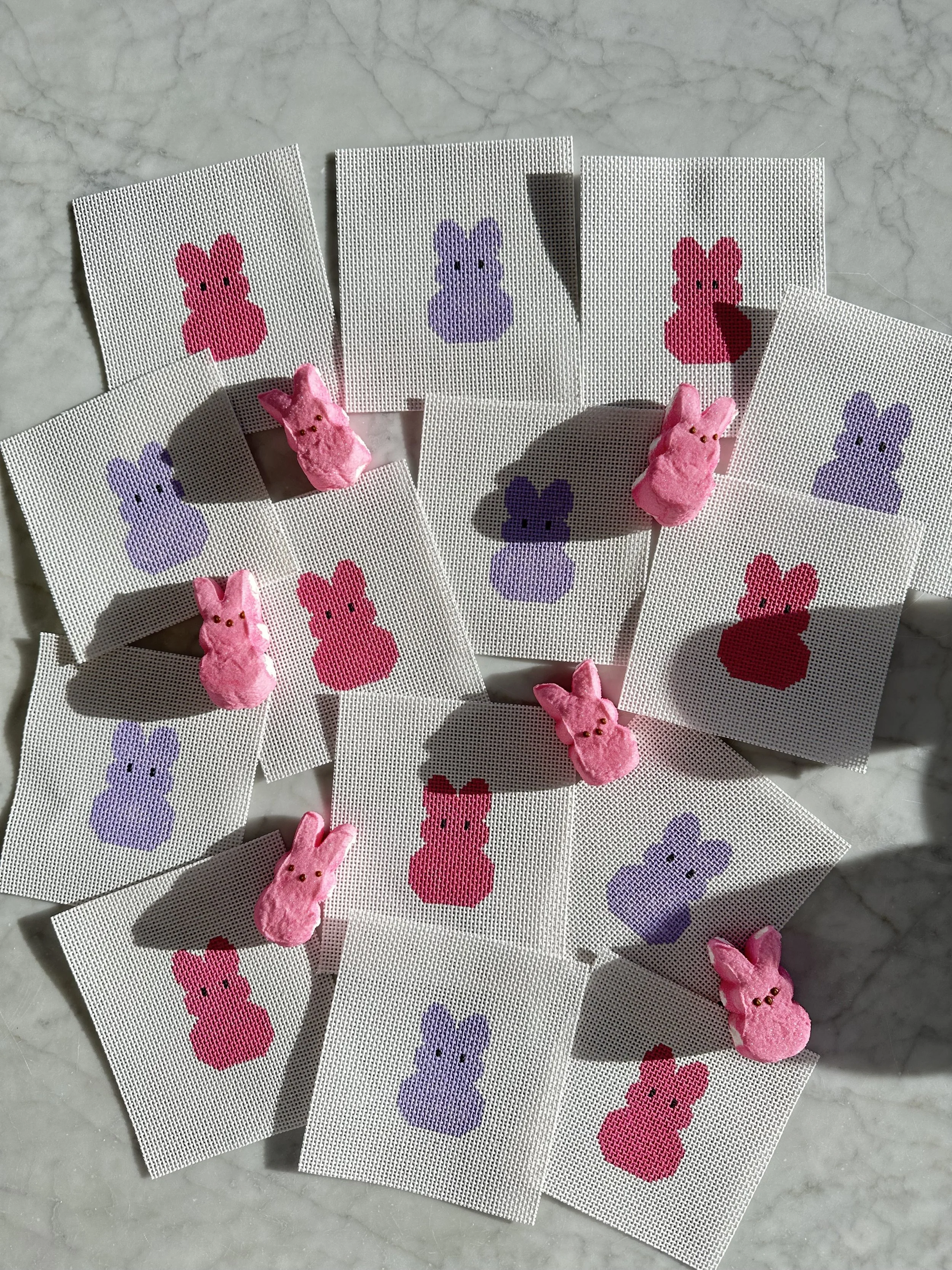 Several cross-stitch cards with colorful bunny designs in pink and purple, with small pink bunny figurines placed on some of the cards, all positioned on a light marble surface.