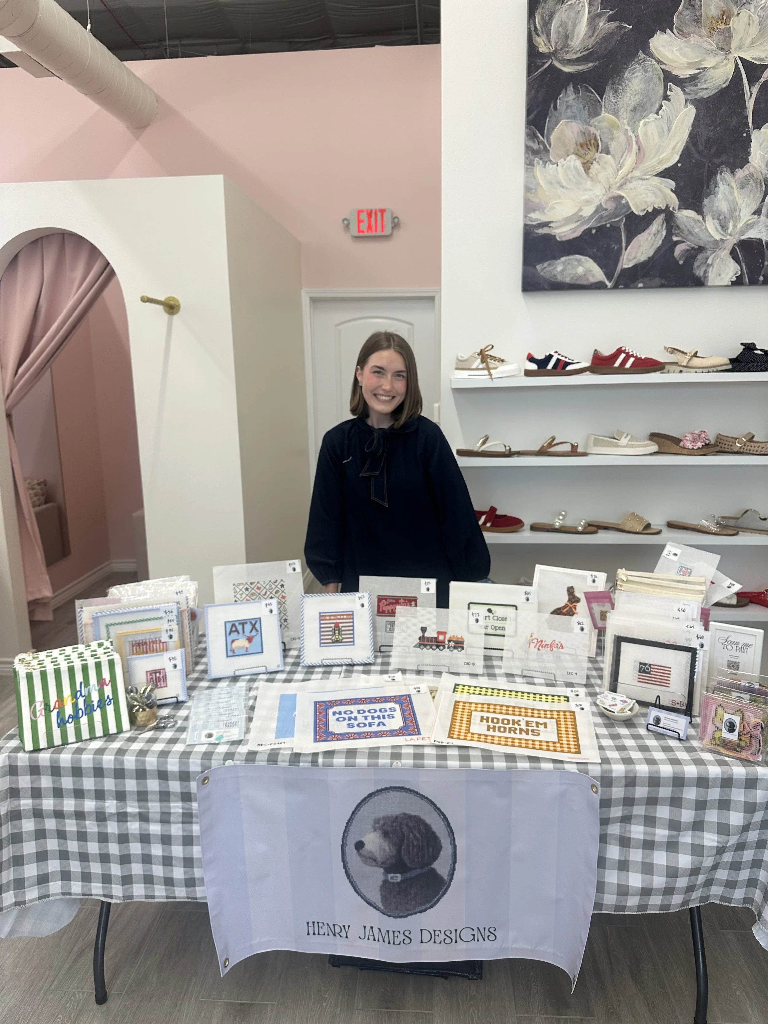 A woman standing behind a table with needlework crafts on display, in a store or craft fair setting, with shelves of shoes and a large floral painting on the wall behind her, and a banner that reads "Henry James Designs" in front of the table.